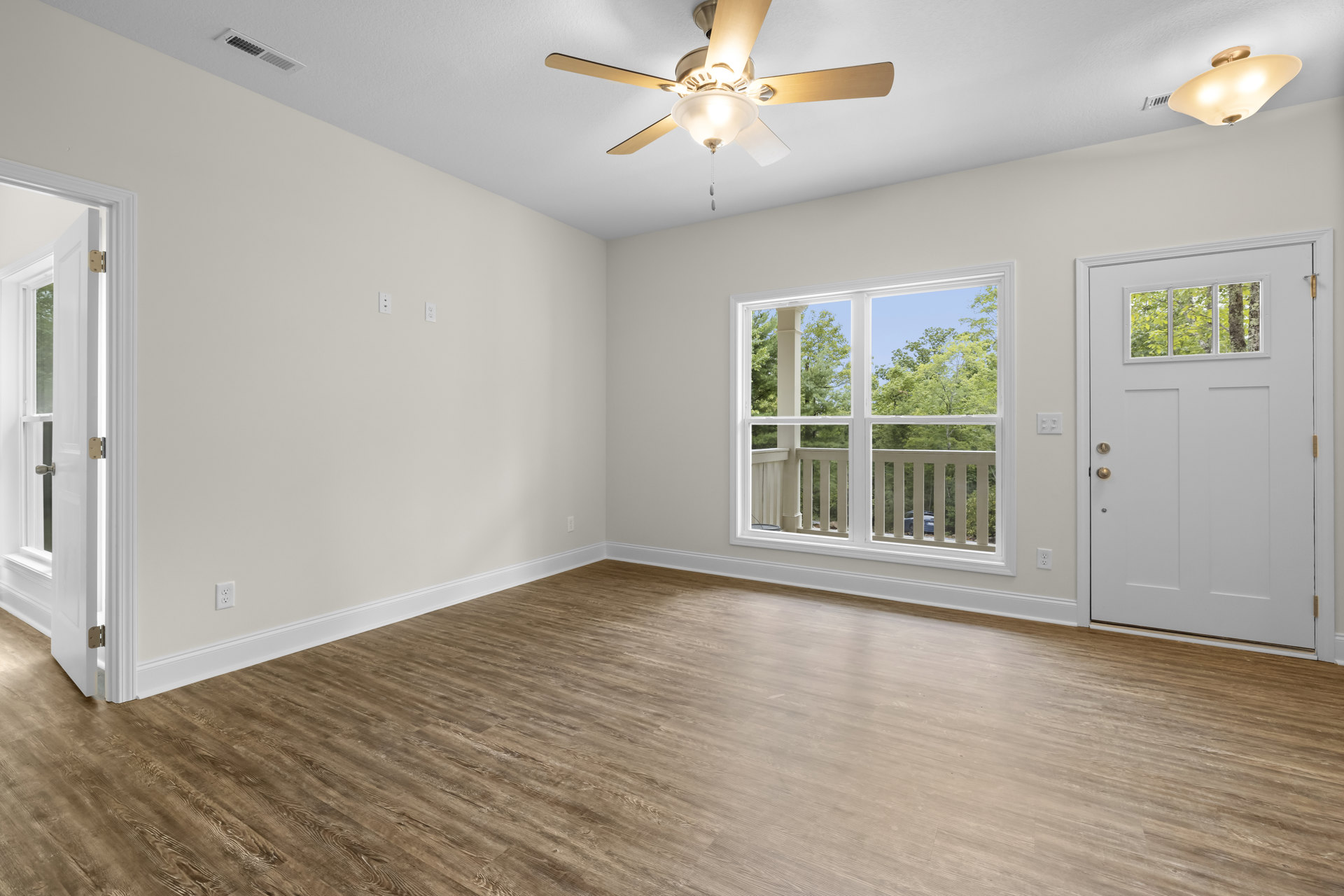 Wood flooring in a bright room with a ceiling fan and light fixture, white door with glass panel, large window overlooking trees.