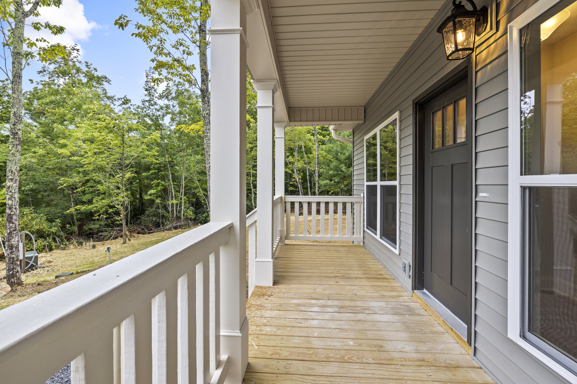 Covered porch with white railing, wooden door, wall-mounted light fixture, grassy yard, trees and mossy trunk in background