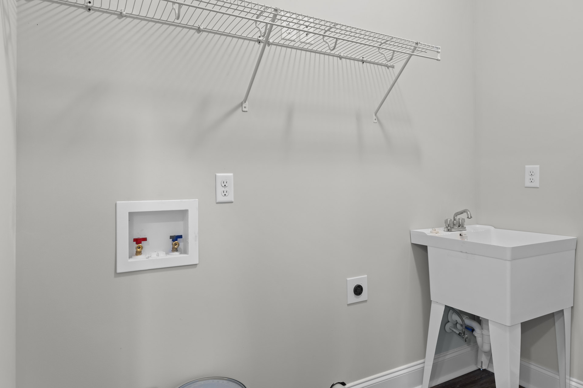 White wall-mounted shelf above a white bathroom sink with exposed plumbing, electrical outlet, and black round outlet on the adjacent surface.