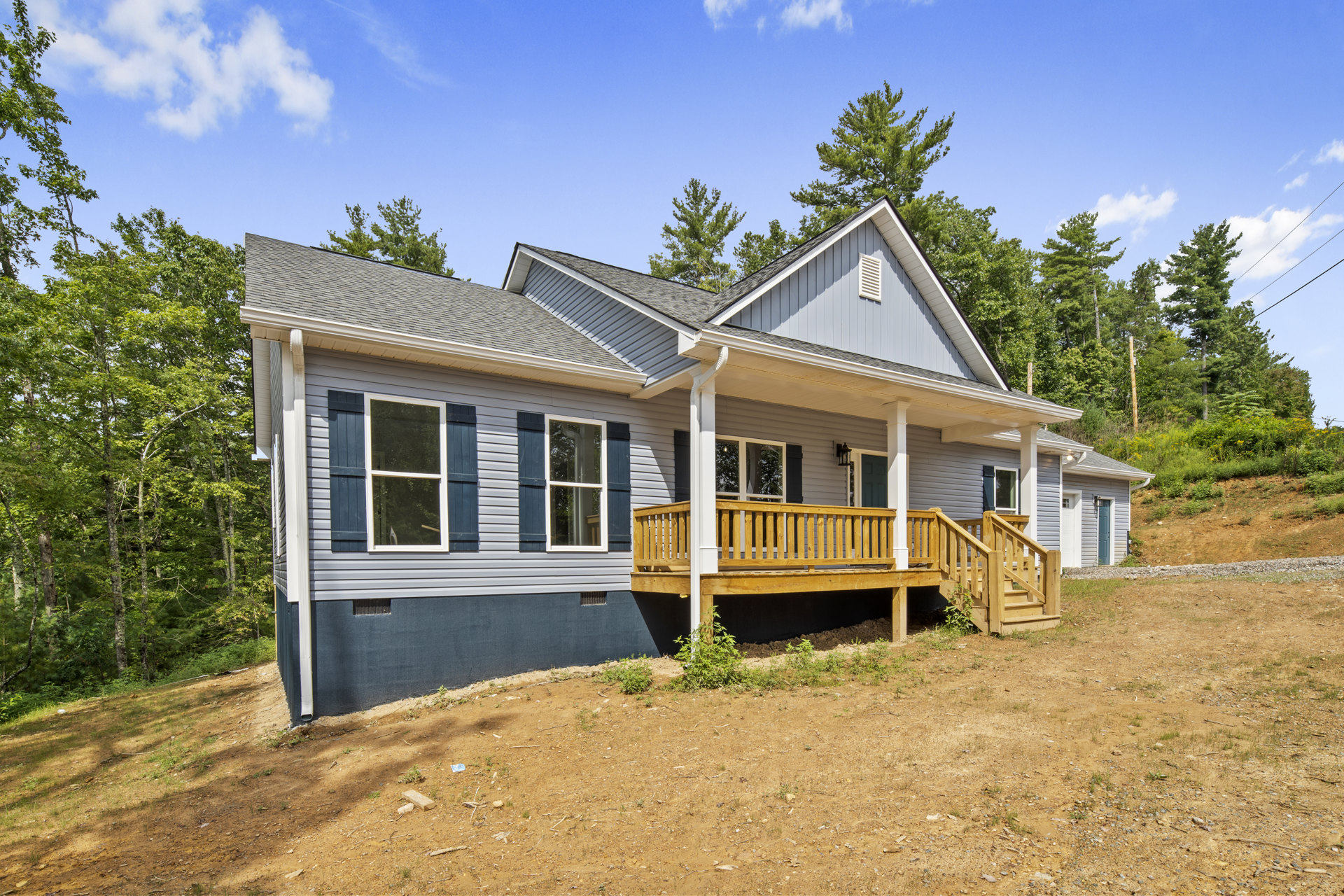 Two-story house with white siding, blue shutters, covered front porch, and wood deck overlooking a dirt patch, surrounded by trees and plants under a cloudy sky