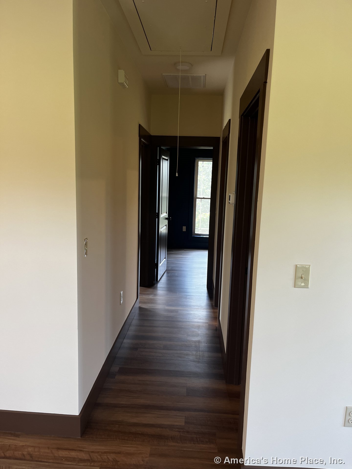 Hallway featuring wood-look plank flooring, dark baseboards and door trim, white walls, ceiling attic access, interior doors, and a view into an adjacent room with a large window.