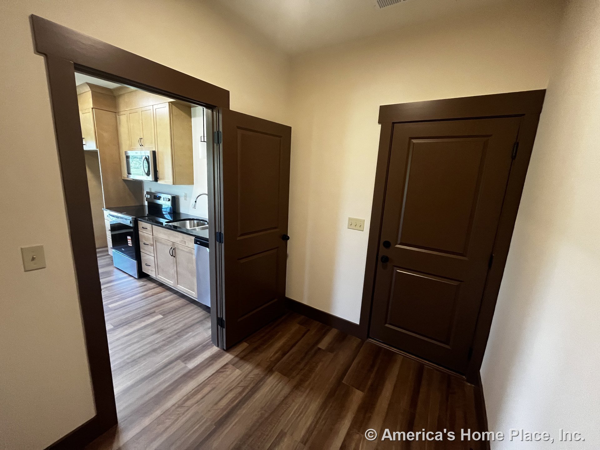 Light wood kitchen cabinetry and stainless steel appliances visible from hallway with brown interior doors, wood-look flooring, and brown trim around doorways.