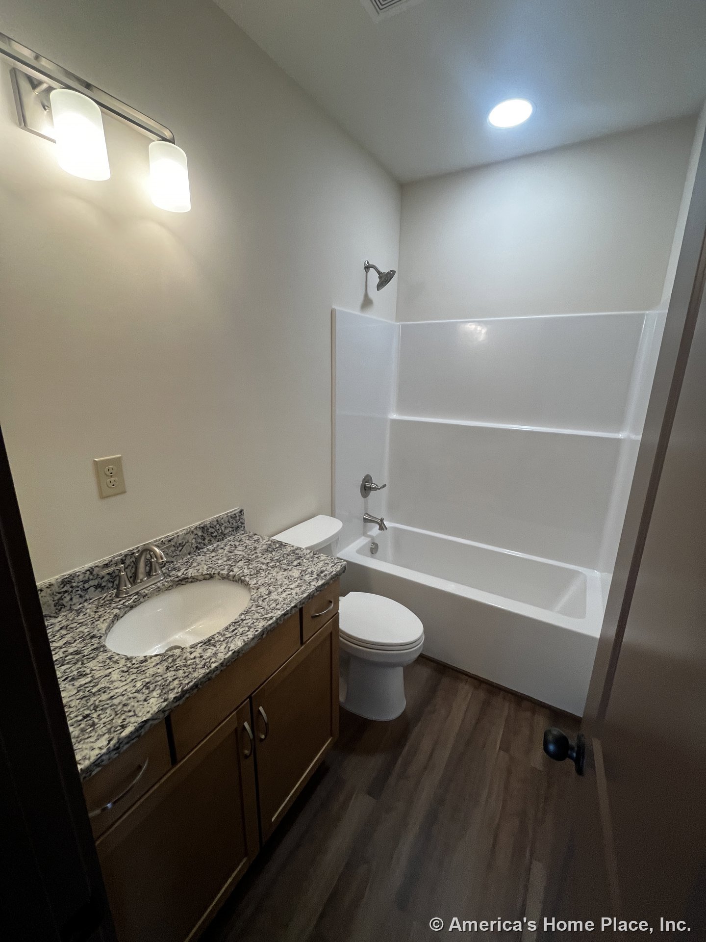 Granite countertop vanity with single sink and chrome faucet, built-in bathtub and shower, wood-look vinyl flooring, wall-mounted light fixture, recessed ceiling light, and