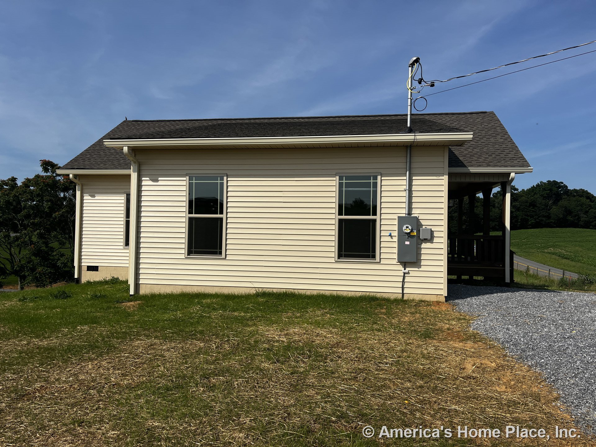 Beige vinyl siding exterior with two double-hung windows, covered porch, asphalt shingle roof, gravel driveway, and visible electrical meter and panel.