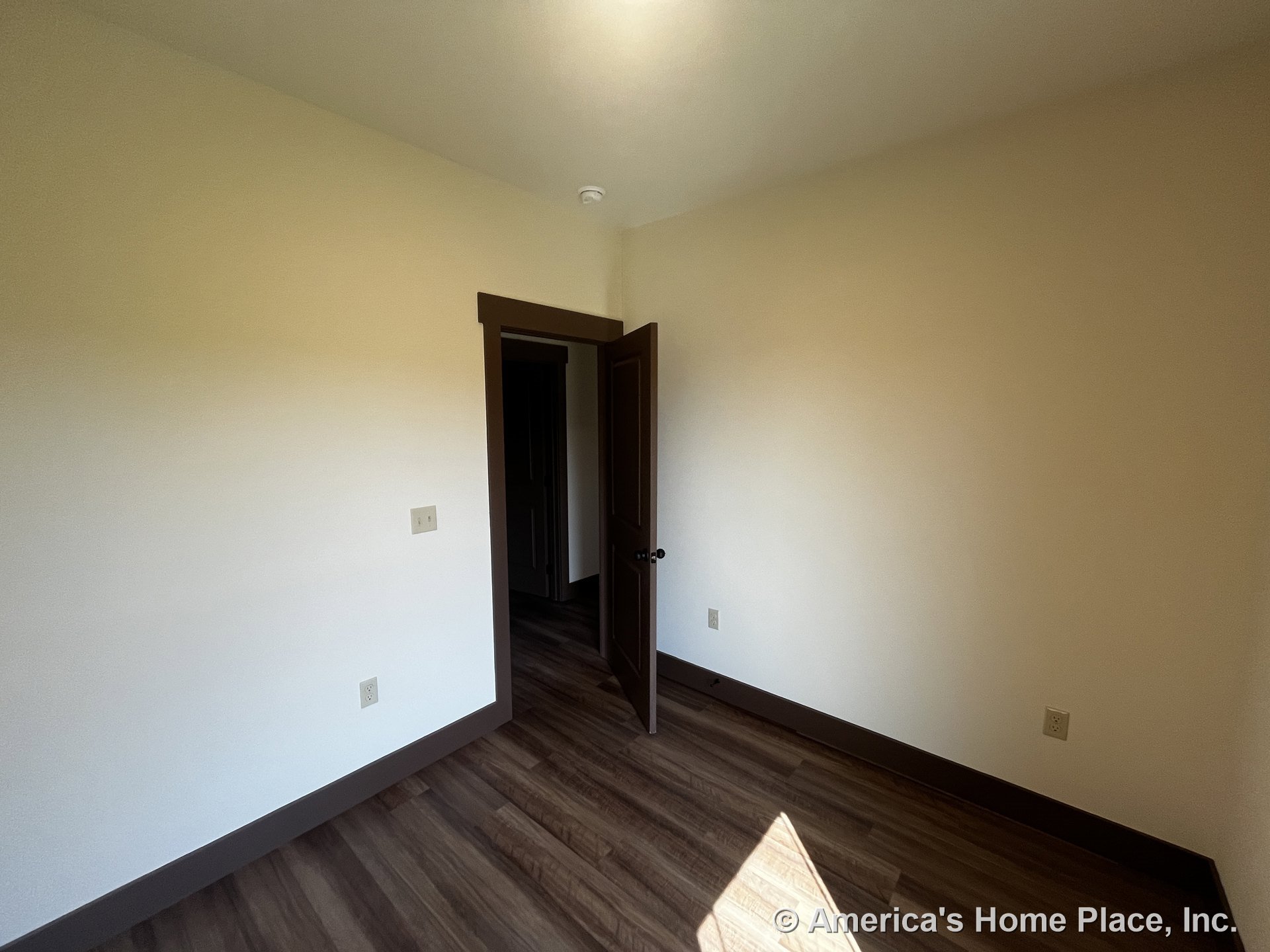 Small interior bedroom with wood-look plank flooring, dark baseboards and door trim, open door, neutral painted walls, ceiling-mounted light fixture, and multiple electrical