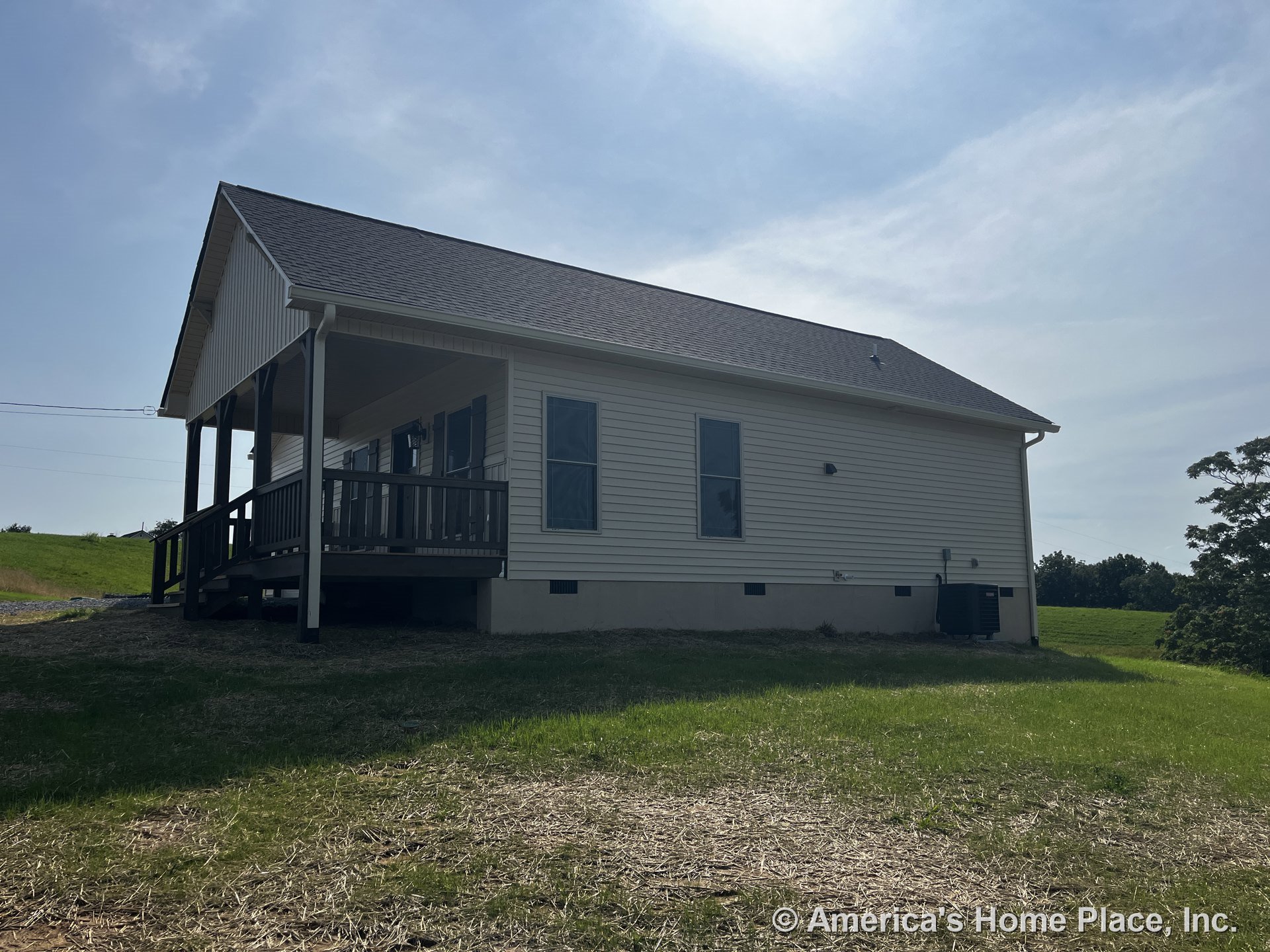 Covered front porch with wood railing, vinyl siding exterior, gabled roof, double-hung windows, exterior HVAC unit, outdoor lighting, gutters, and rural setting.