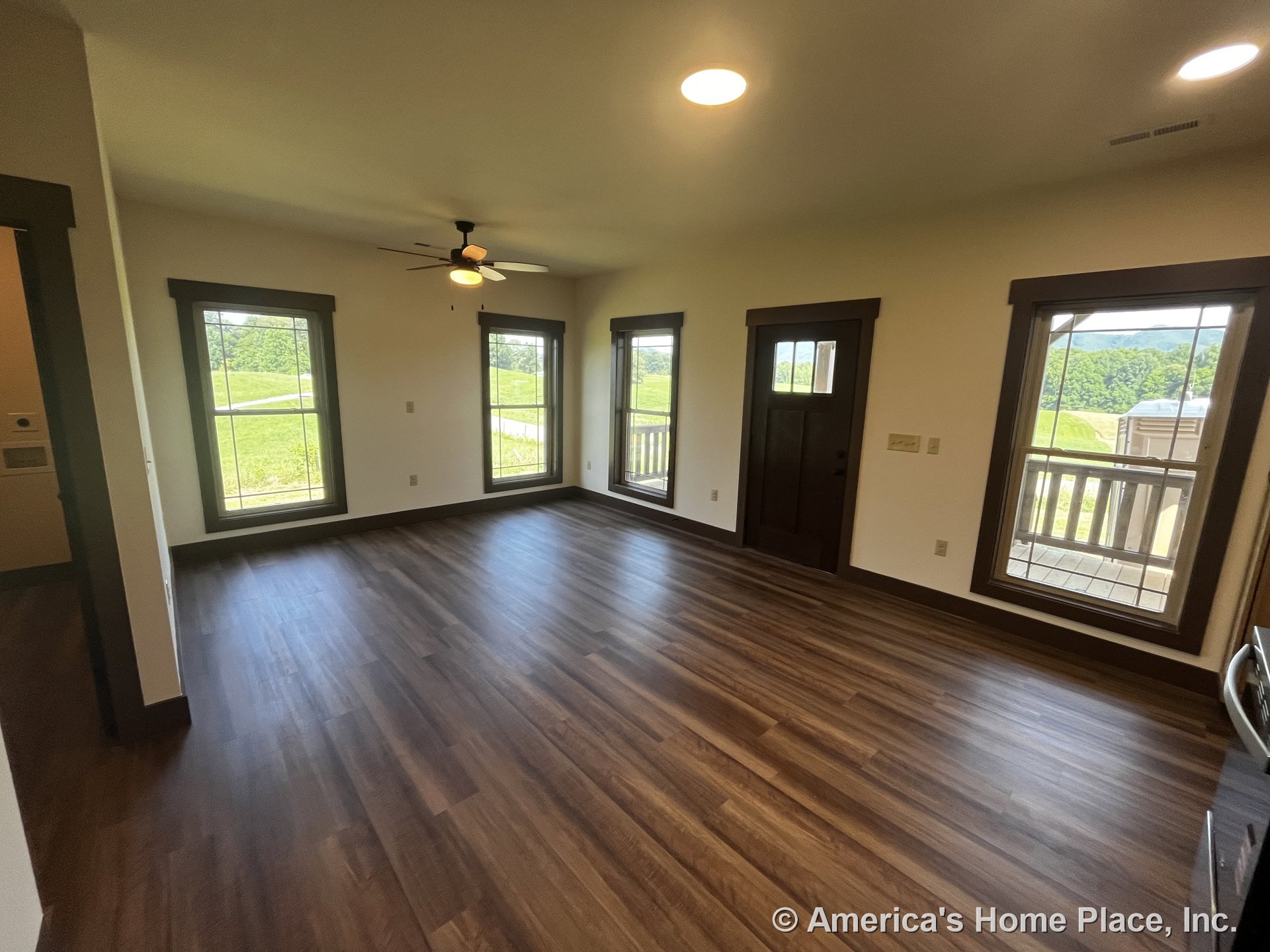Living room with three tall windows framed by dark wood trim, vinyl plank flooring, ceiling fan with light, recessed ceiling lights, exterior door with glass panel, neutral walls