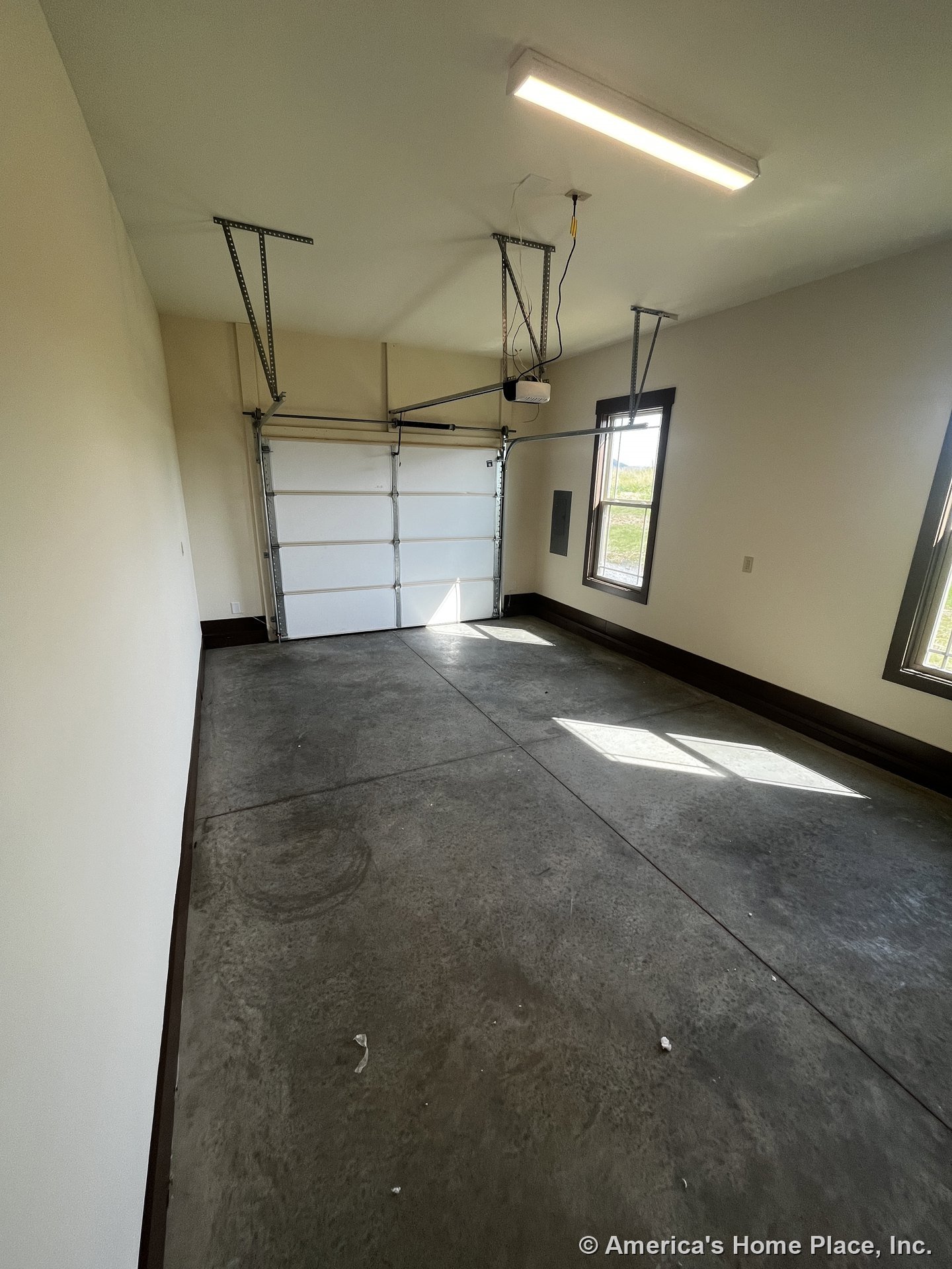 Garage interior with concrete slab flooring, overhead garage door, rectangular window with trim, ceiling-mounted light fixture, wall-mounted electrical panel, and dark baseboard
