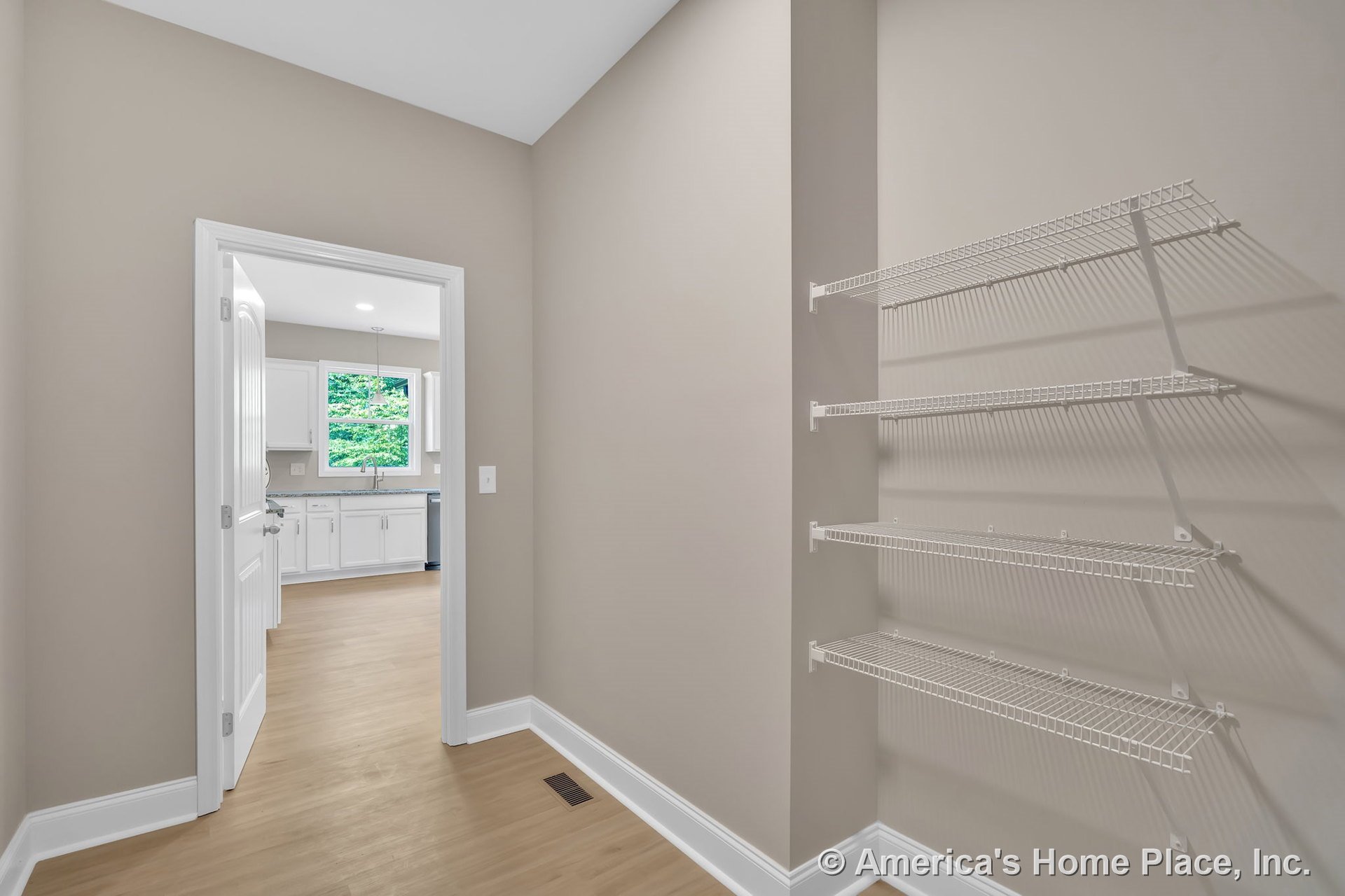 Pantry with built-in wire shelving next to kitchen with white cabinetry, large window, light wood flooring, white trim, and door.