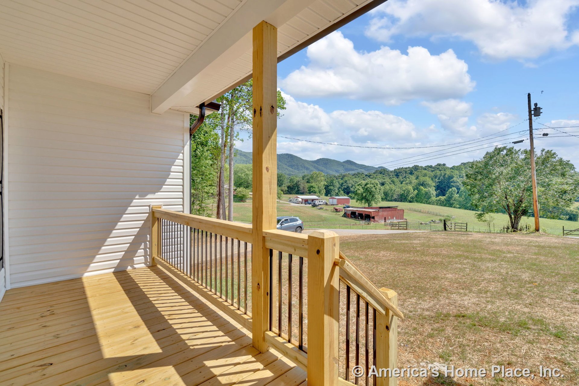 Covered front porch with wood decking, vertical wood posts, metal balusters, white vinyl siding, beaded ceiling, and open rural landscape beyond railing.