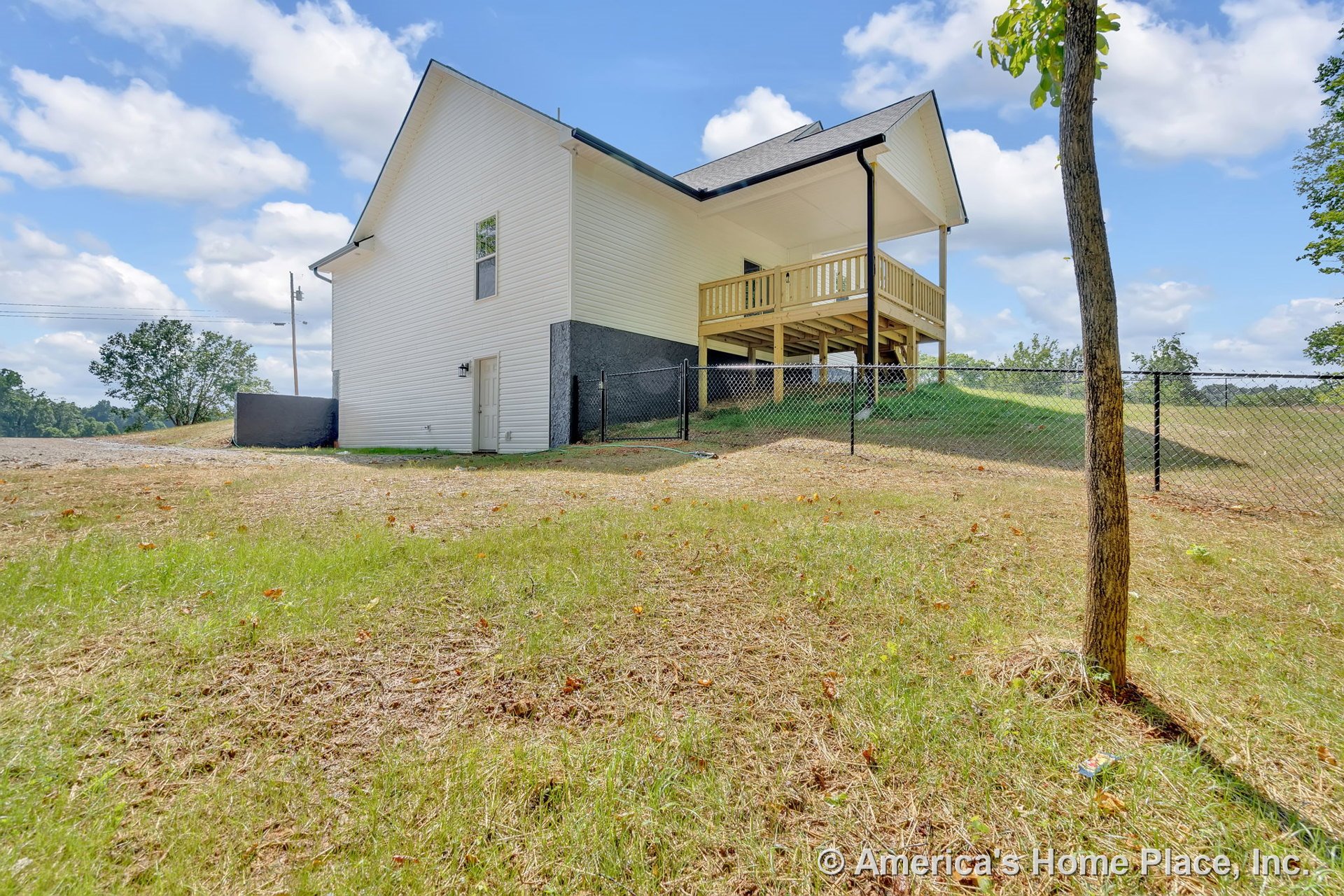 White vinyl siding exterior with covered elevated porch featuring wood railings, single exterior door, black chain-link fence enclosing sloped backyard, multiple windows, roof