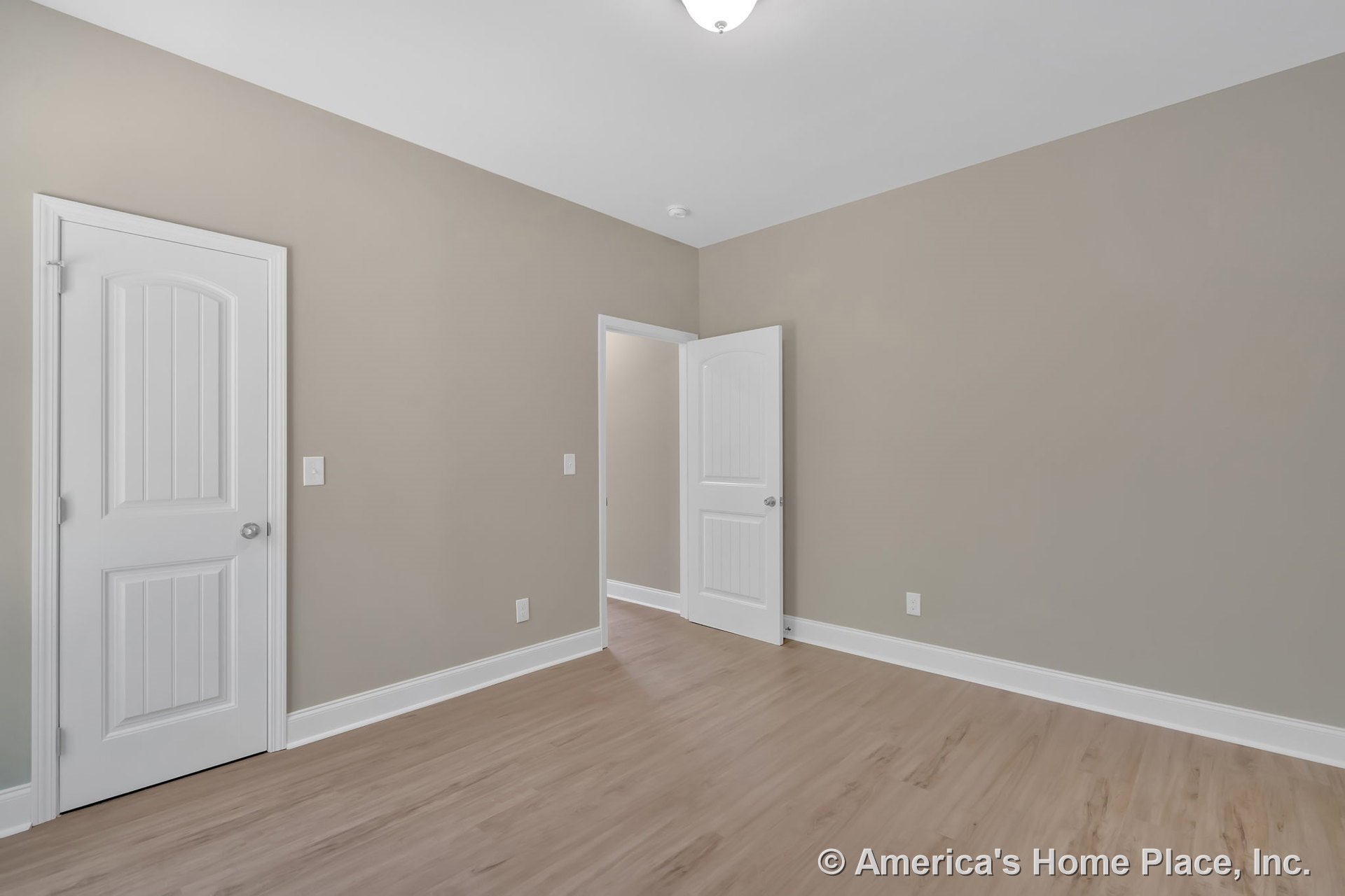 Bedroom with light wood flooring, beige walls, white baseboards, white paneled doors, flush mount ceiling light, and minimalist trim details.