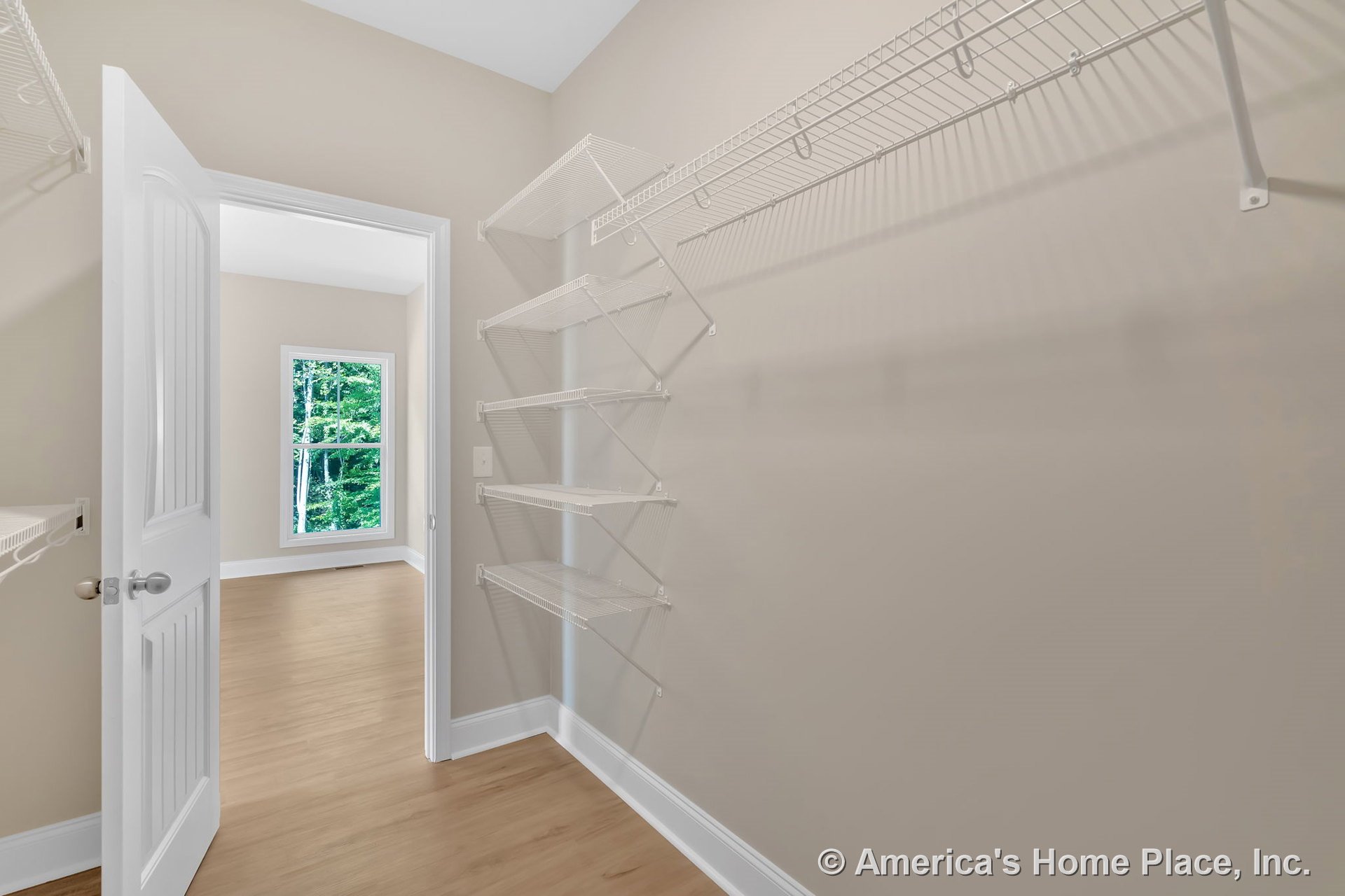Walk-in closet with built-in white wire shelving and hanging racks, open door, light wood flooring, trimmed window, and neutral wall color.