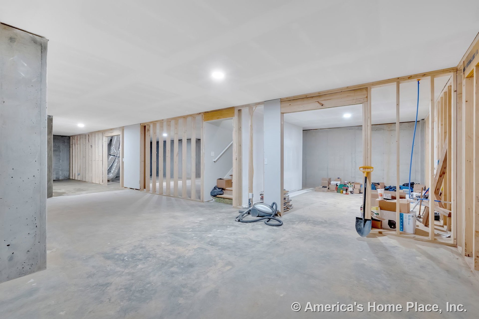 Unfinished basement with exposed wood framing, concrete floors, open stairway, recessed ceiling lights, unfinished drywall ceiling, and visible utility area.