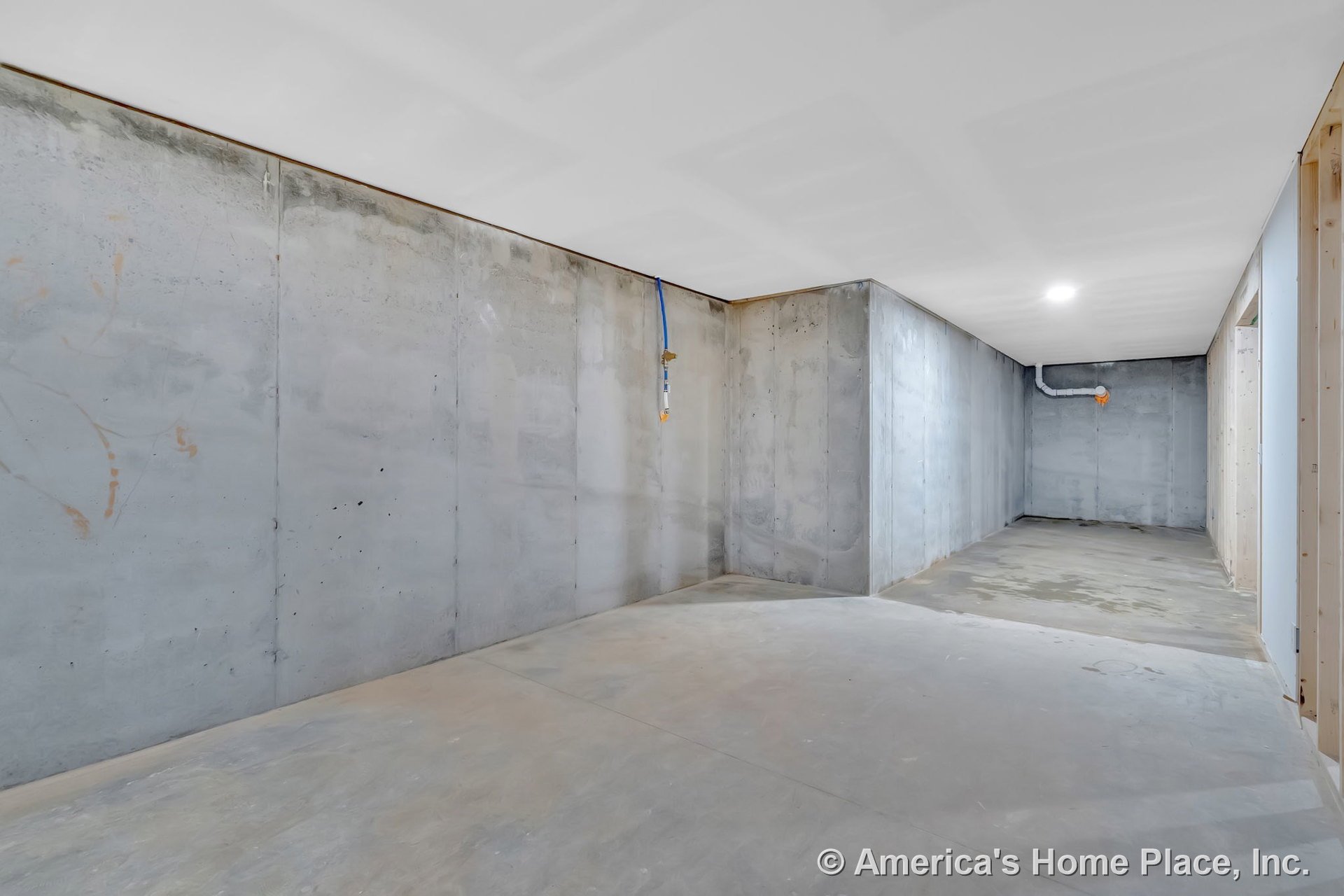 Unfinished basement with concrete walls and floor, exposed plumbing pipes, recessed ceiling lights set in drywall, and wood framing along the right side.
