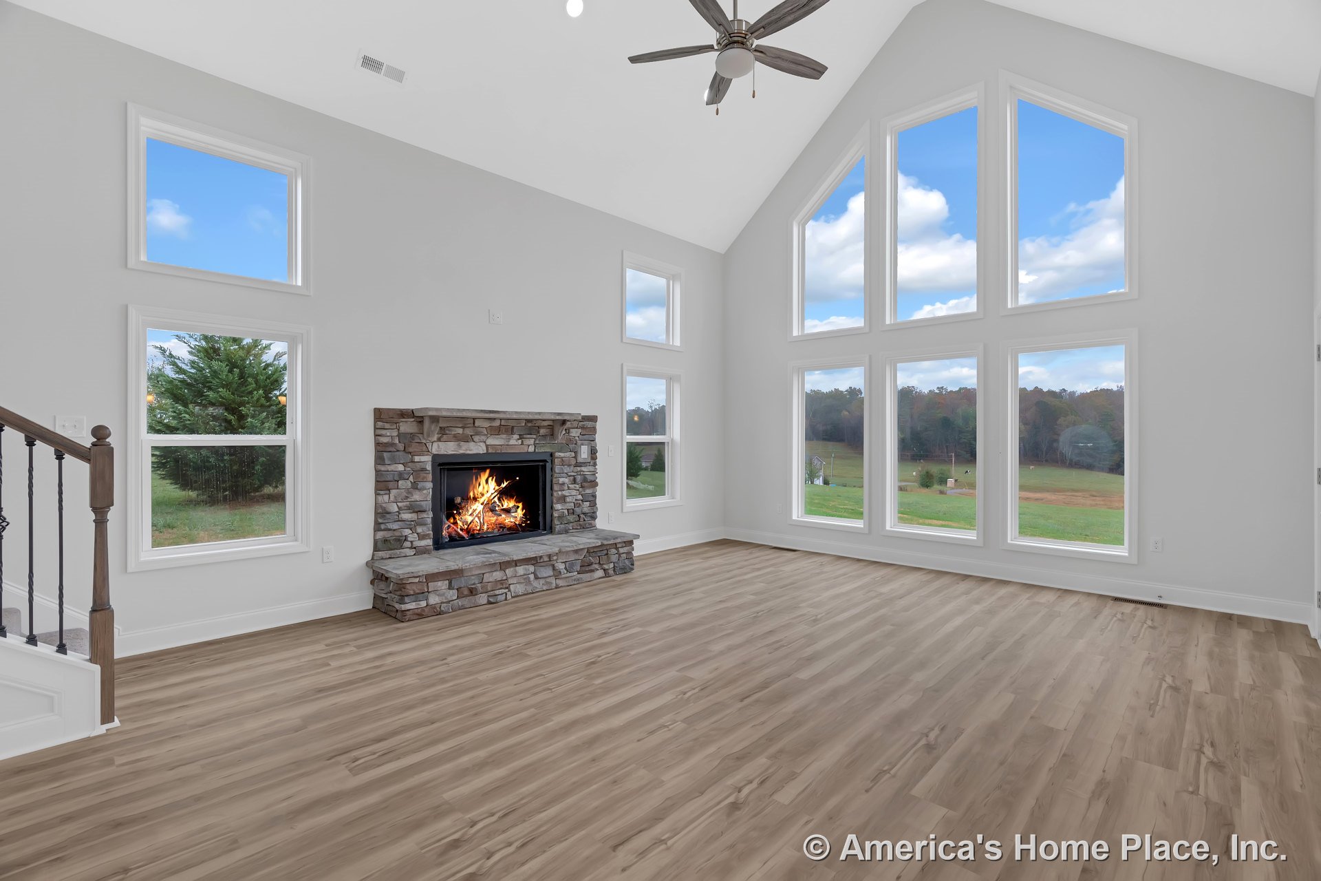 Vaulted ceiling living room with large picture windows, stone fireplace, wood-look flooring, white trim, ceiling fan, and open stair railing.