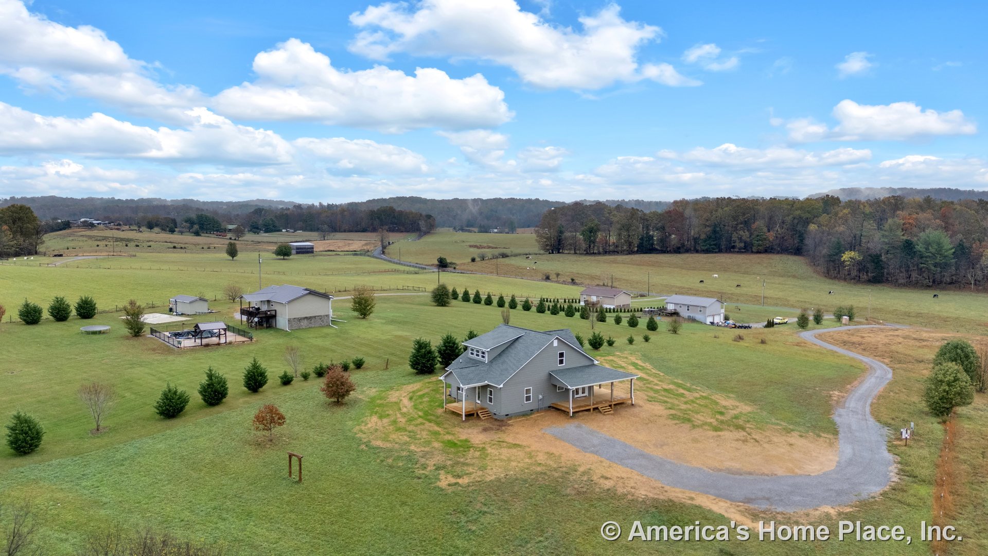 Gray two-story home with wood trim, covered front porch, dormer windows, gravel driveway, expansive yard, and rural landscape.