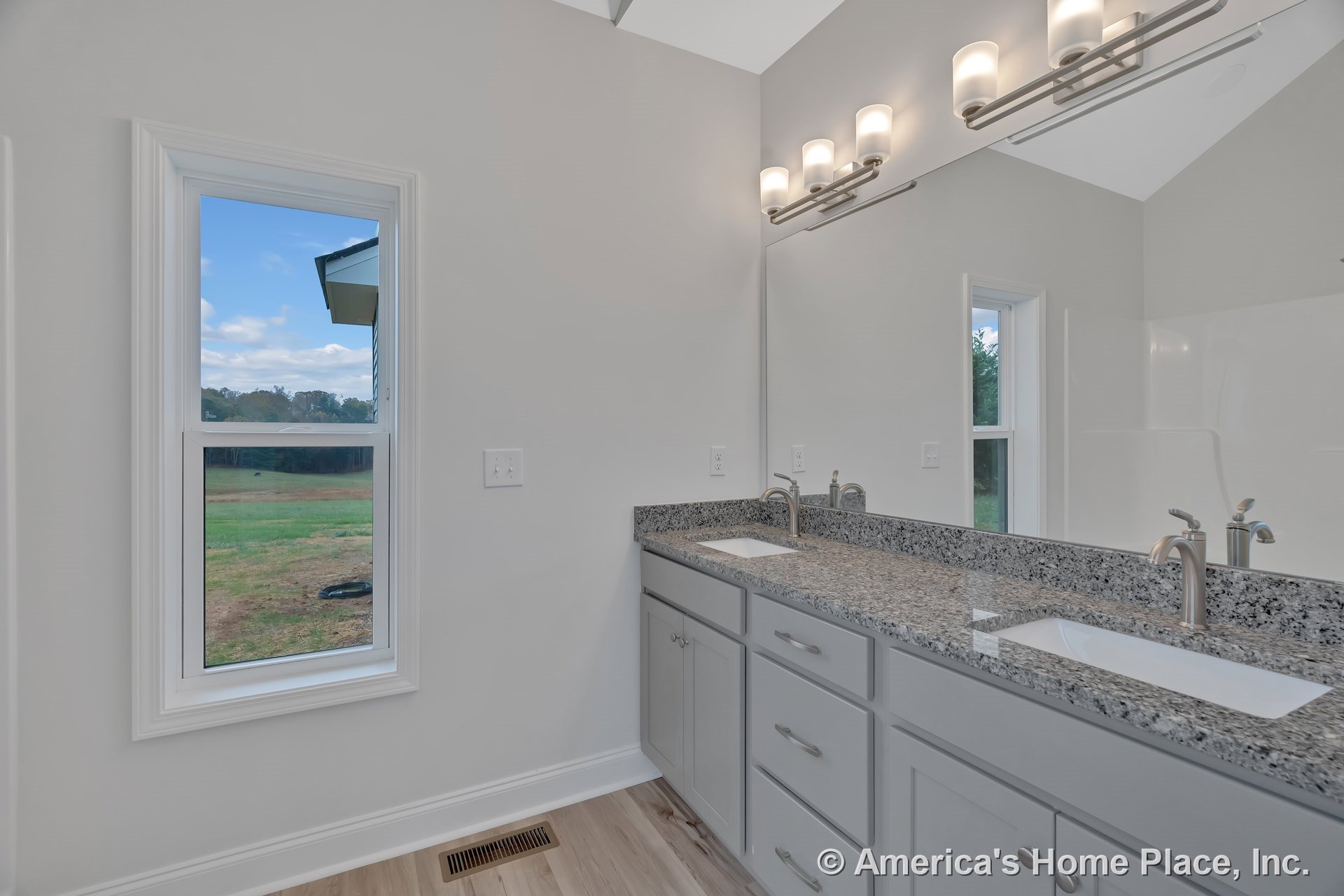 Double vanity with granite countertop, gray shaker cabinets, two undermount sinks, large wall mirror, modern chrome vanity lighting, white trim window, light wood flooring, and