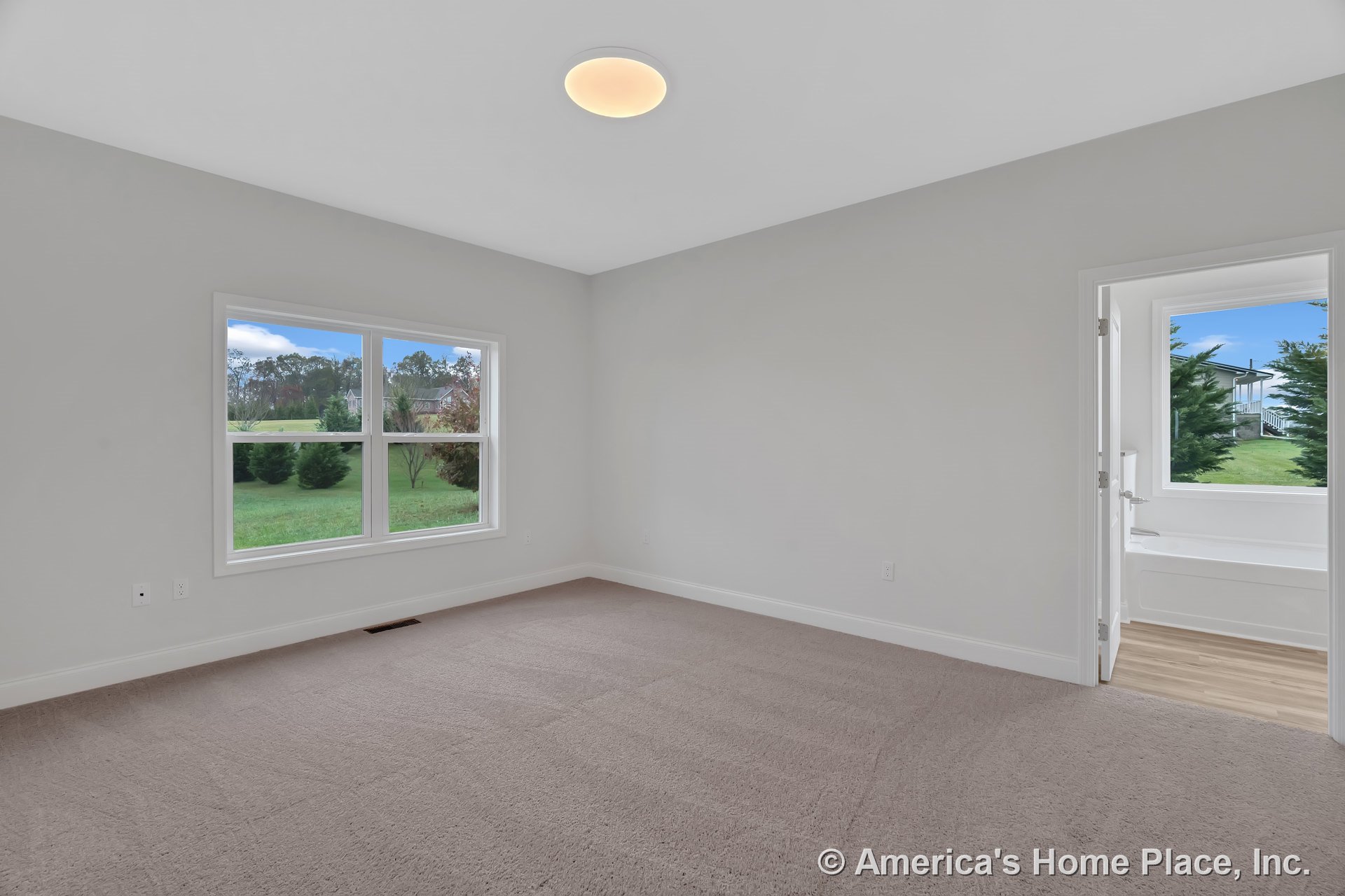 Carpeted bedroom with large double window, white baseboards, flush ceiling light, and doorway leading to bathroom featuring wood-look flooring and additional window.