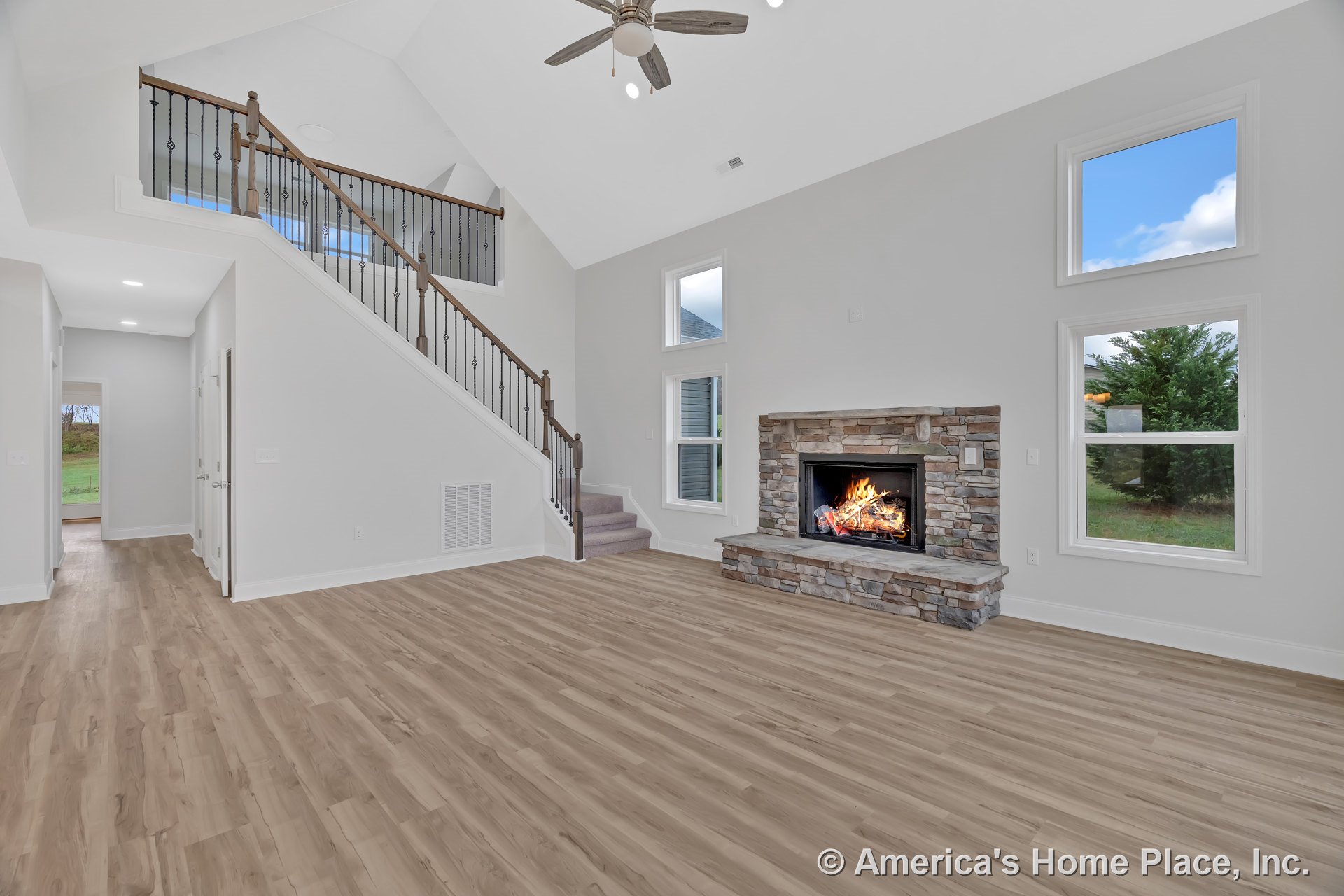 Stone fireplace with hearth, vaulted ceiling and ceiling fan, light wood plank flooring, double stacked windows allowing natural light, open staircase with iron balusters, white