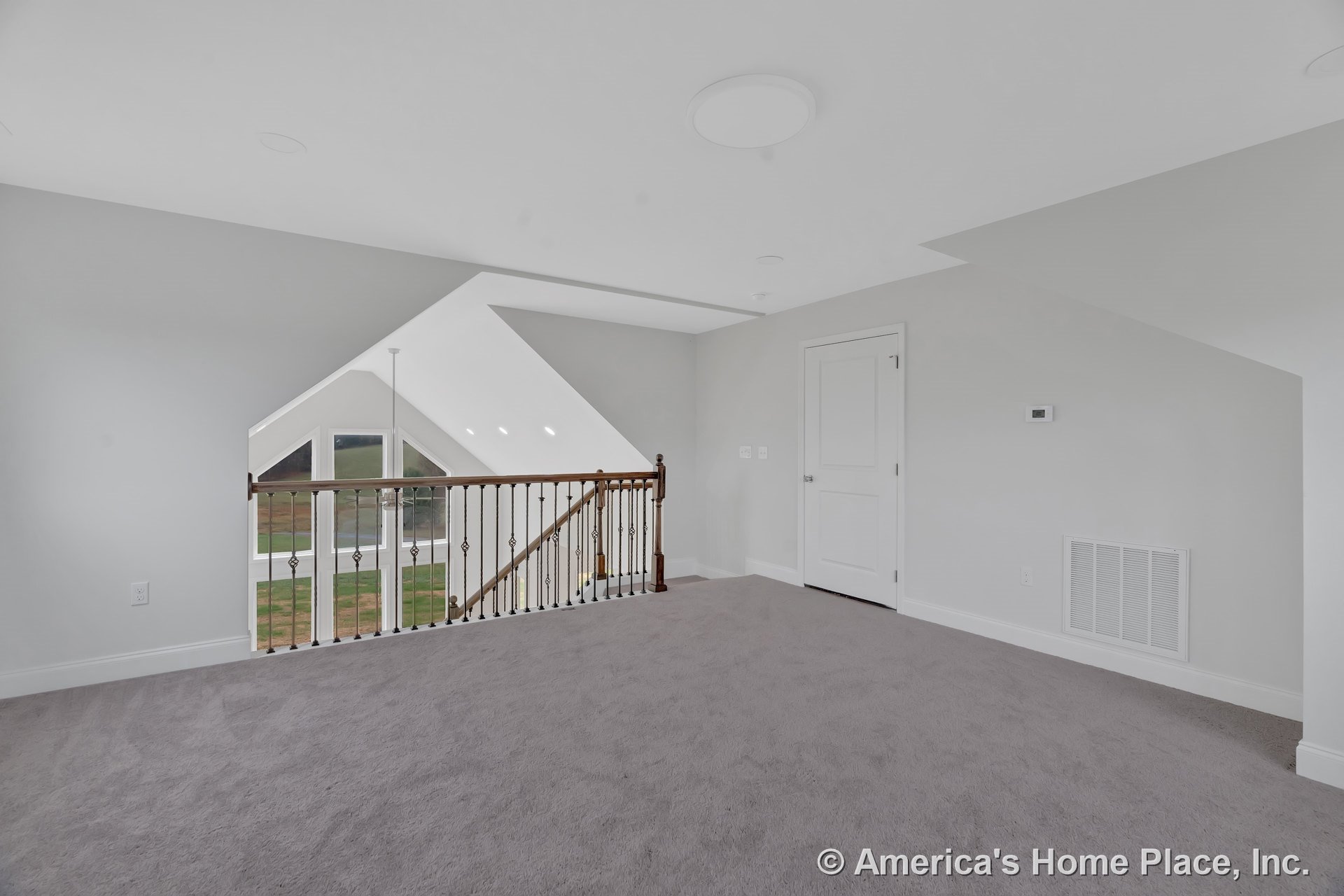 Carpeted loft with wood and iron stair railing, large triangular windows framed by white trim and baseboards, recessed ceiling lighting, and paneled interior door.
