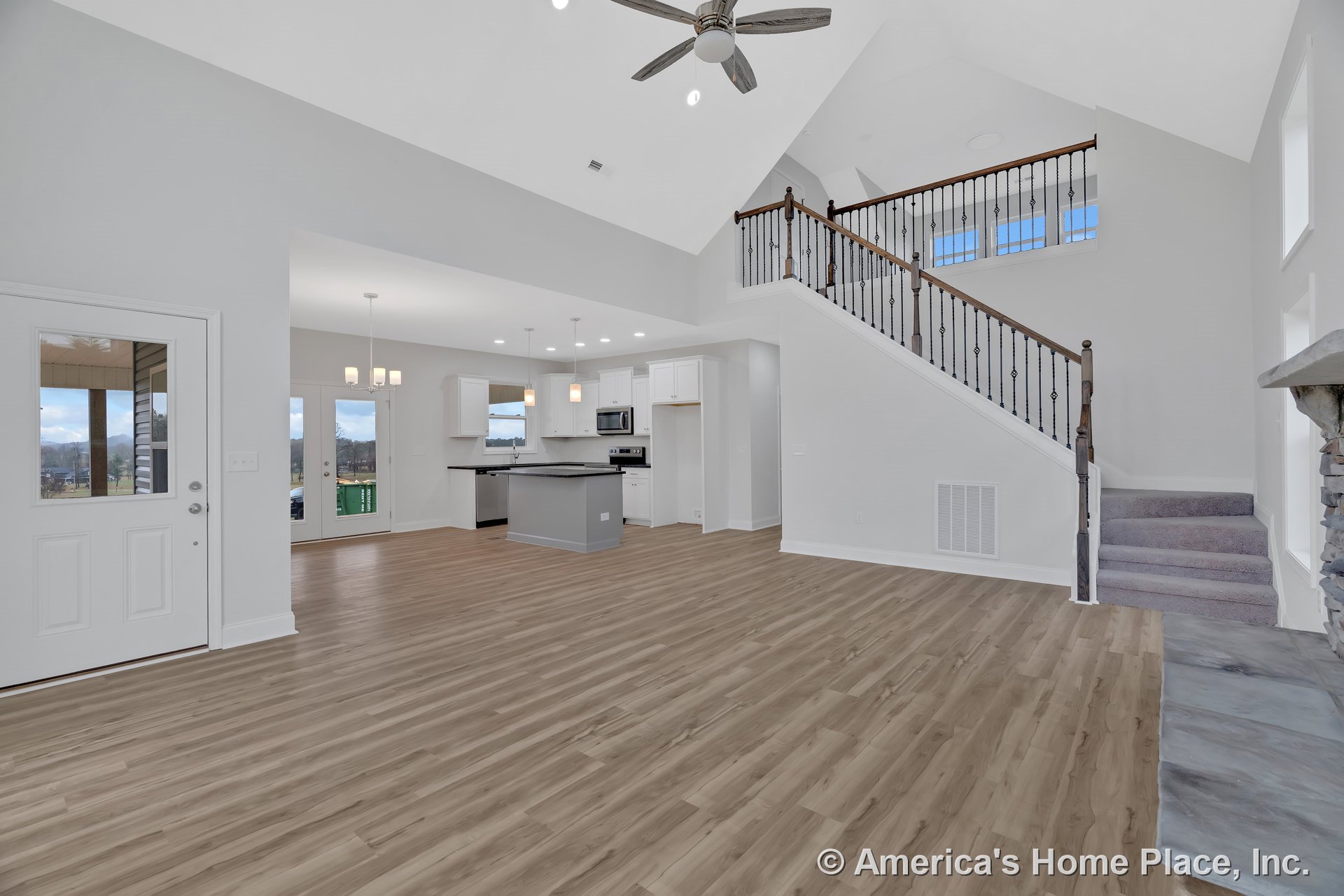 Vaulted ceiling with ceiling fan above open-concept living area, wood-look plank flooring throughout, iron and wood staircase railing, white kitchen cabinetry, stone fireplace