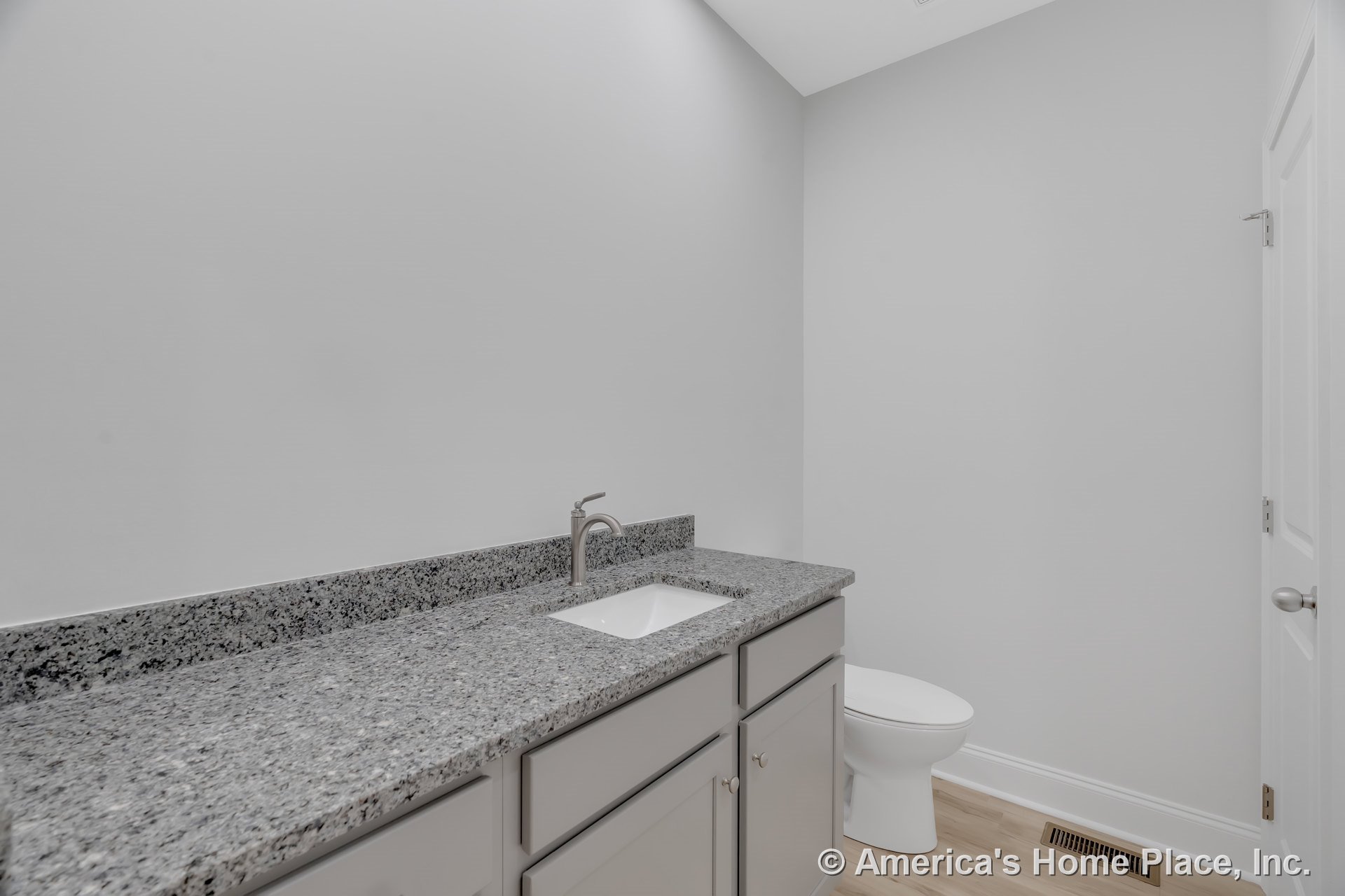 Gray granite countertop with undermount sink atop light gray cabinetry, white toilet adjacent, light wood flooring, white walls and trim, vent cover near door.