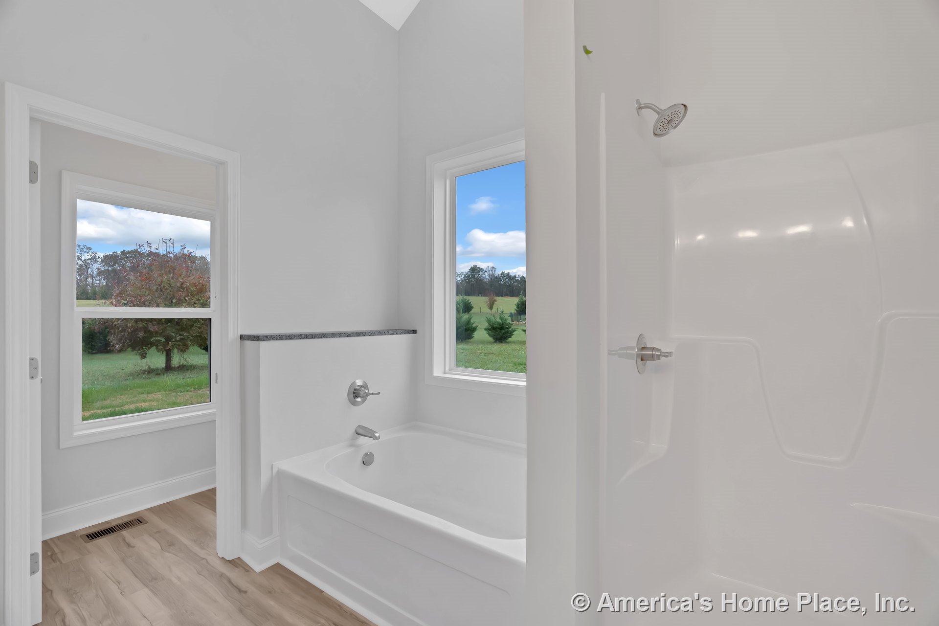 Built-in white bathtub with chrome fixtures beside a separate glass shower enclosure, large windows offering natural light, light wood flooring, white trim and baseboards, and