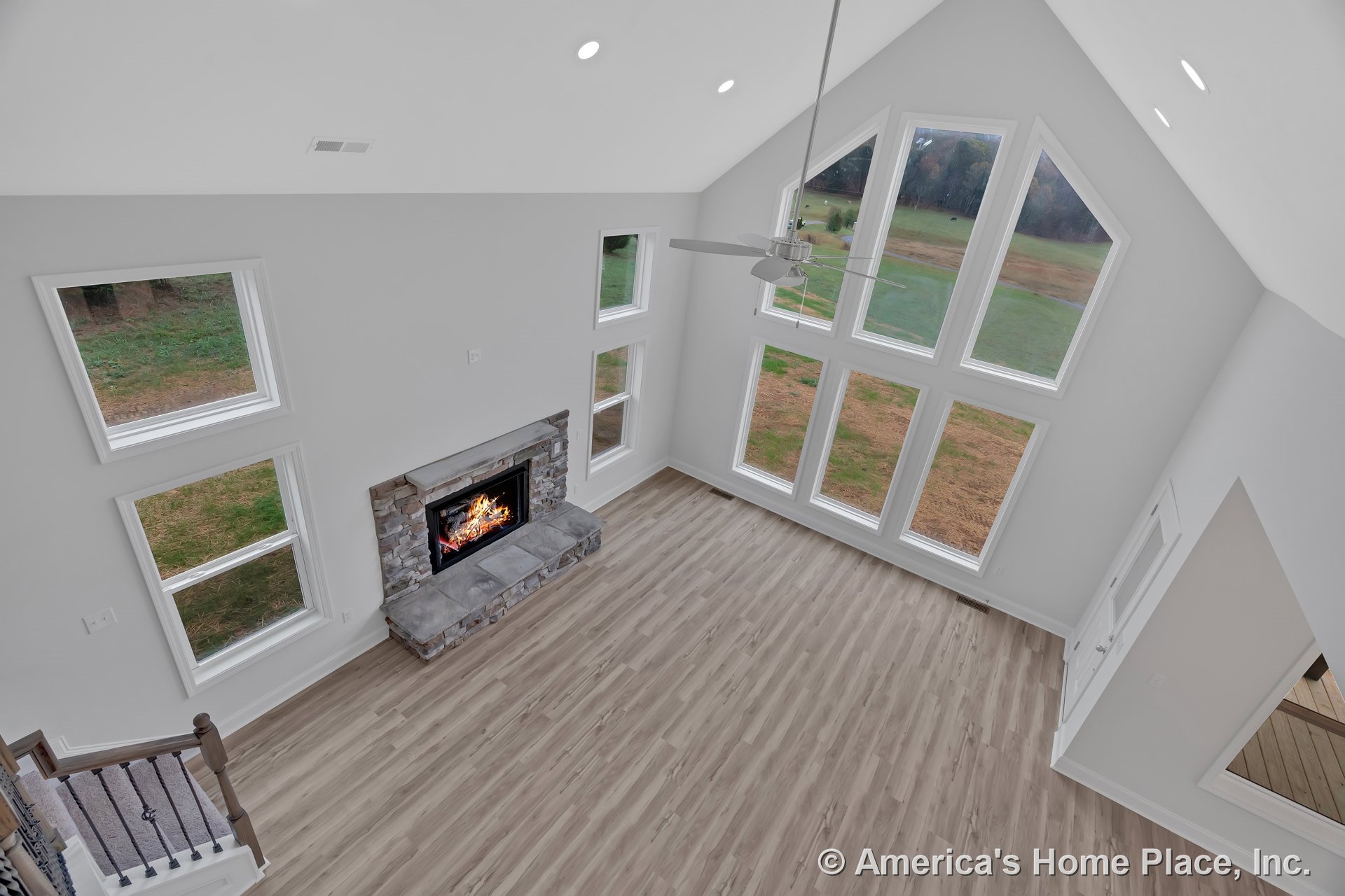 Vaulted ceiling living room with floor-to-ceiling windows, stone fireplace surround, wood-look plank flooring, recessed ceiling lights, white trim and baseboards, open layout, and