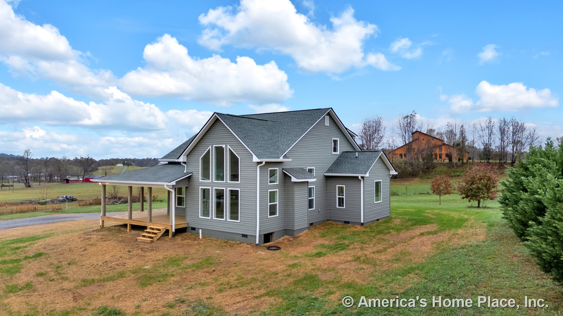 Modern gray vinyl siding exterior with multiple large windows, covered porch with steps, gabled roof, white trim accents, set in a rural landscape.