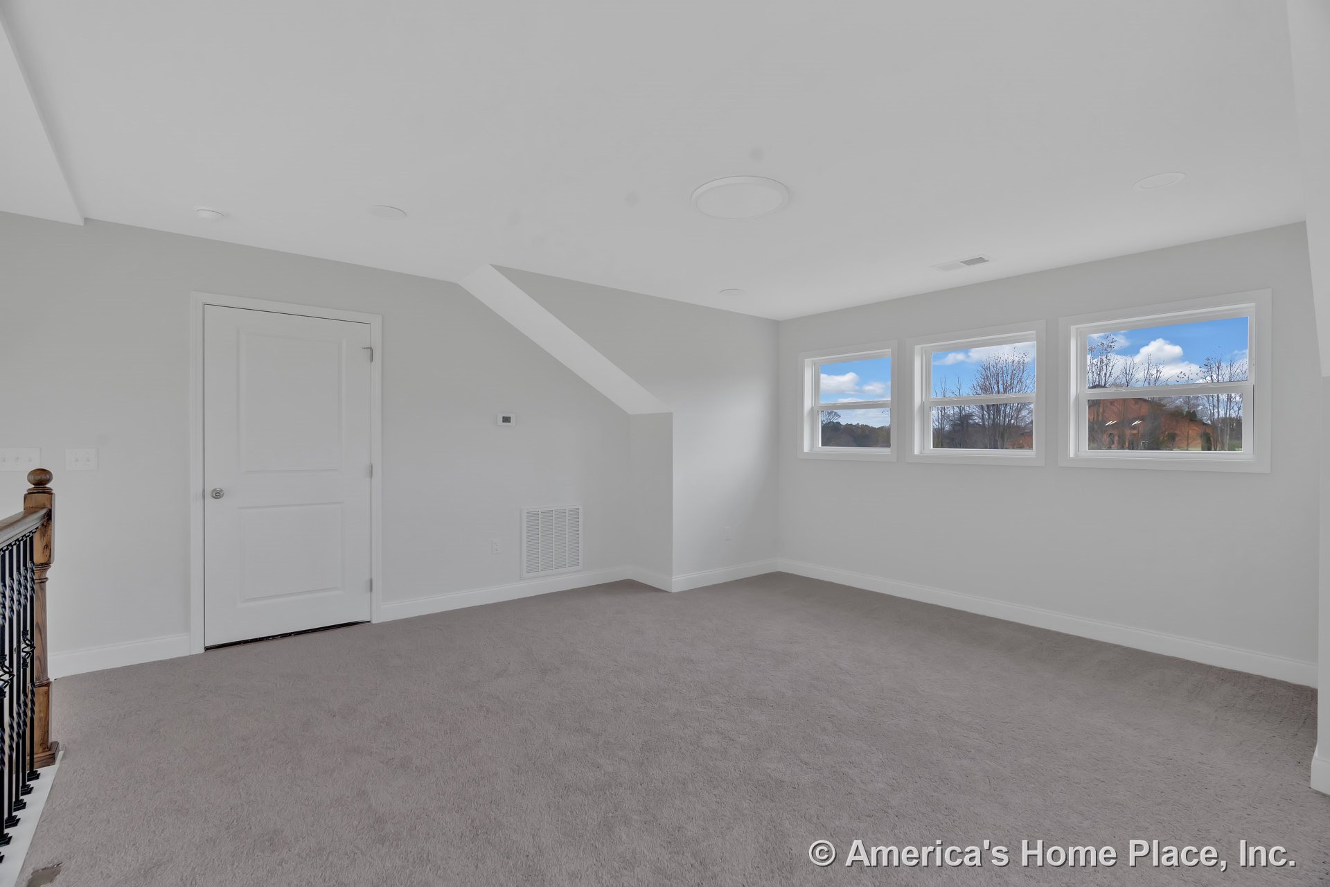Upstairs bonus room with three rectangular windows, neutral carpet flooring, white trim and baseboards, angled ceiling, white paneled door, wood and iron stair railing, vent cover