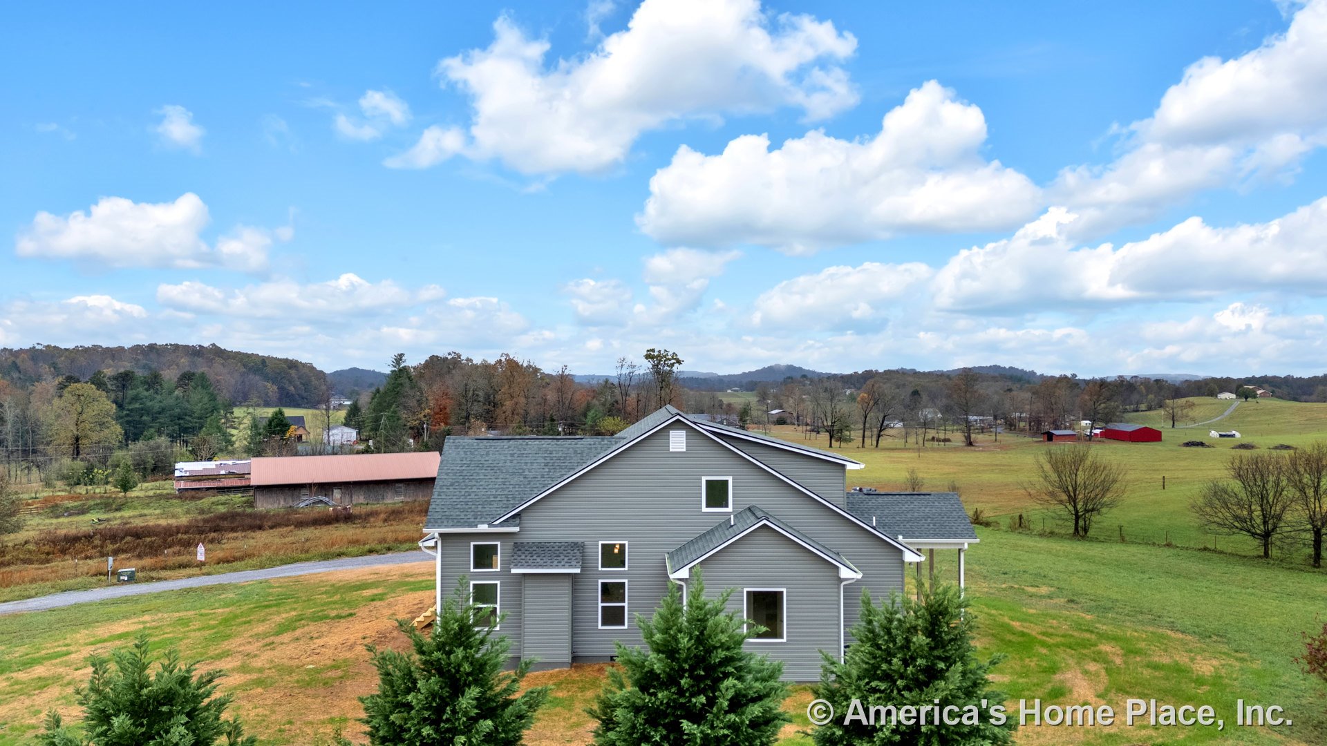 Gray vinyl siding exterior with white trim, asphalt shingle roof, multiple rectangular windows, covered porch entry, and rural landscape backdrop.