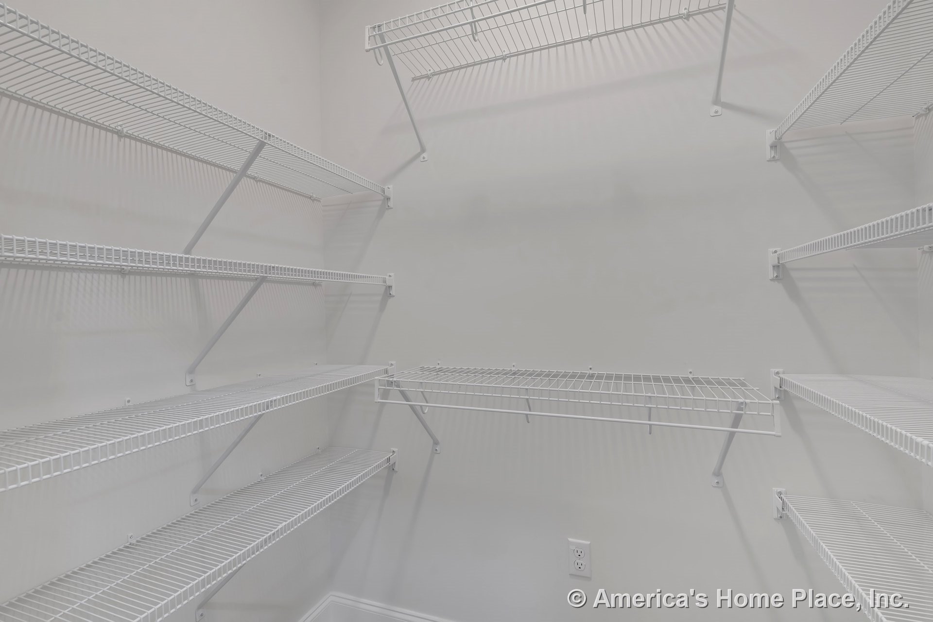 Walk-in pantry with multiple white wire shelving units mounted on light gray walls, baseboard trim, and visible electrical outlet for organized storage.