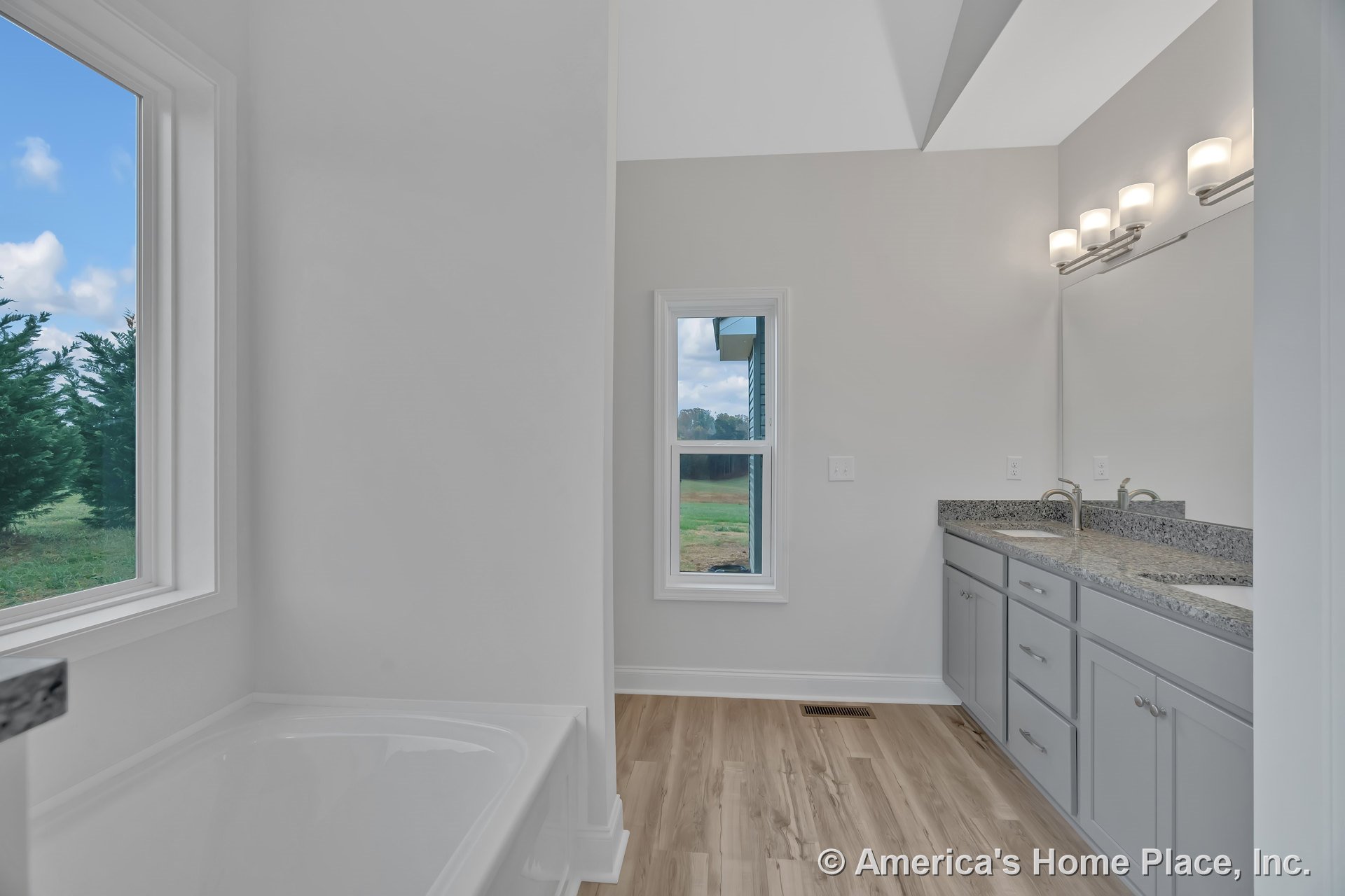 Double vanity with granite countertops and gray cabinetry, soaking tub beside large windows, light wood flooring, wall-mounted vanity lighting, expansive mirror, neutral walls, and