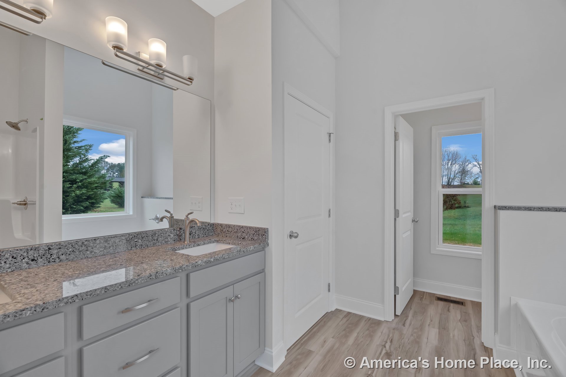 Double vanity with granite countertops and gray shaker cabinets, wall-mounted light fixtures above a large mirror, wood-look flooring, neutral painted walls, window and door with