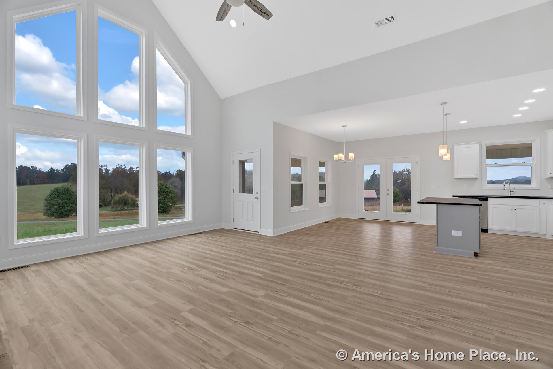 Vaulted ceiling with ceiling fan above open living area, floor-to-ceiling windows along one wall, modern kitchen with white shaker cabinets, black countertops, pendant lighting