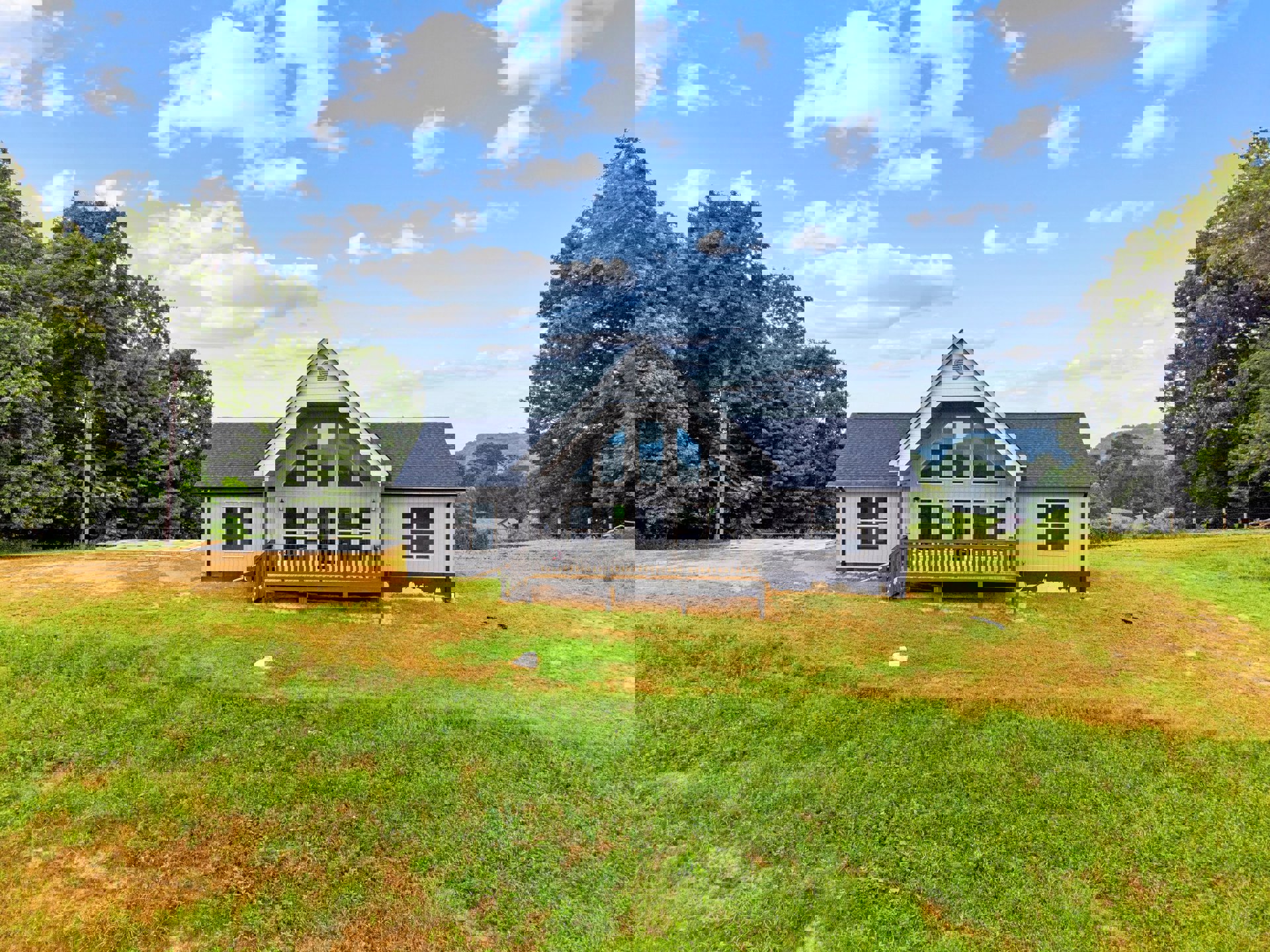 White farmhouse with covered porch, wood deck with railing, grassy lawn, leafy tree, and blue sky with scattered clouds