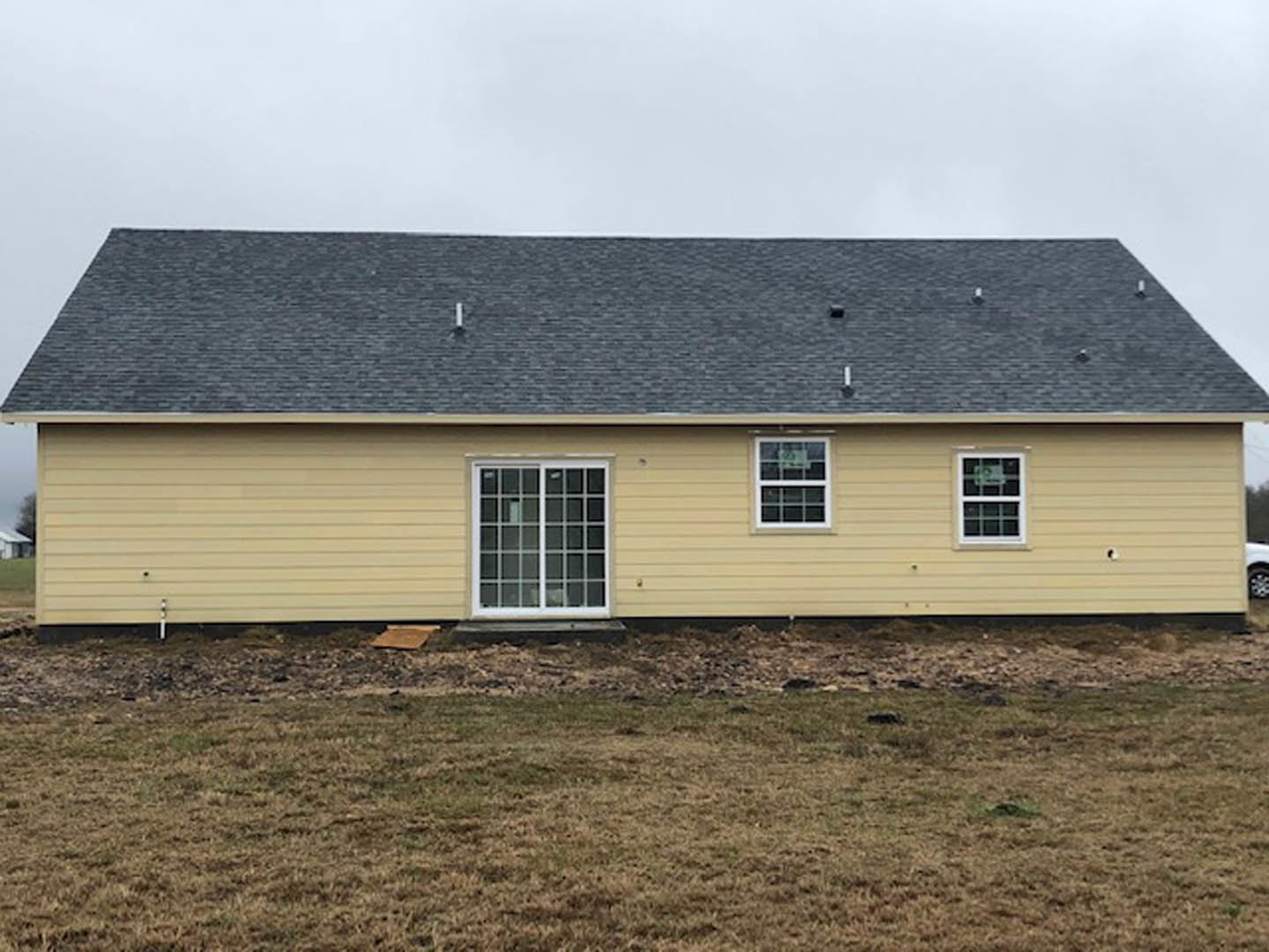 Two-story cottage-style home with gray siding, multi-pane windows, and gabled roof, set on a green lawn with a person standing in the distance under a partly cloudy sky