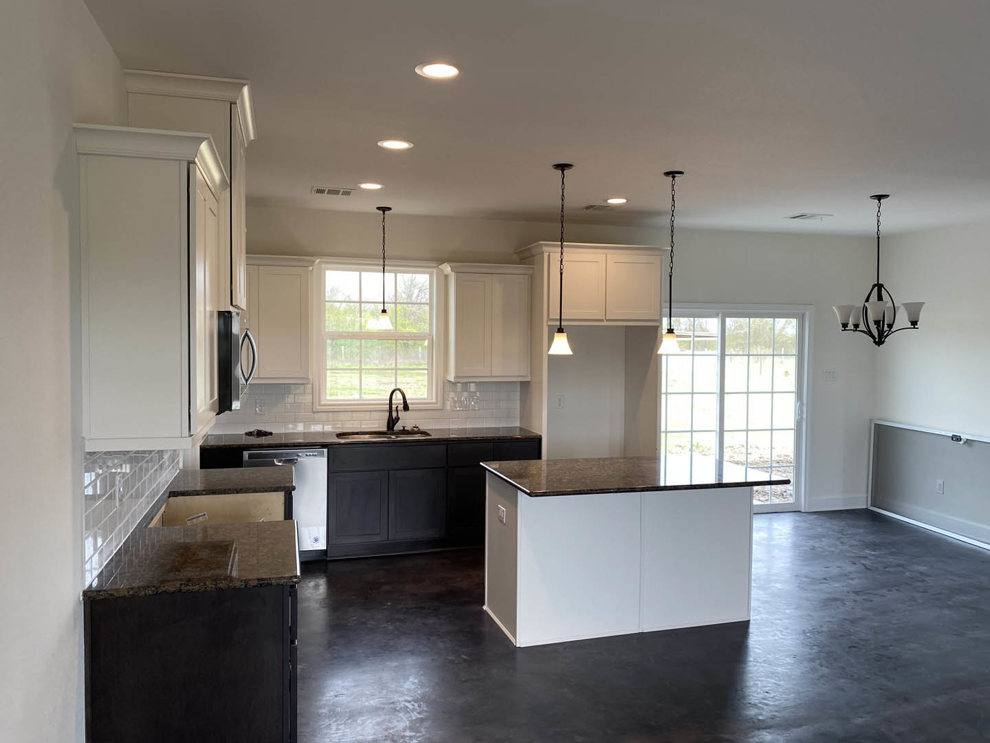 Black stone countertop with white shaker cabinets, stainless steel sink, pendant chandelier with white glass shades, light wood flooring, and neutral painted walls