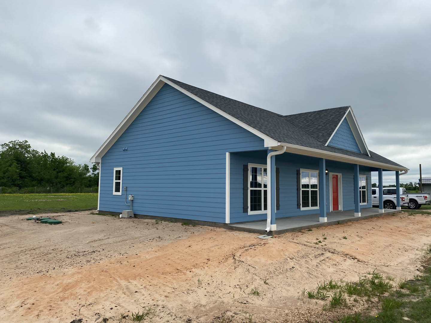 Blue house with white roof, dirt road in front, cluster of trees in background, white truck parked nearby, close-up window visible on exterior.