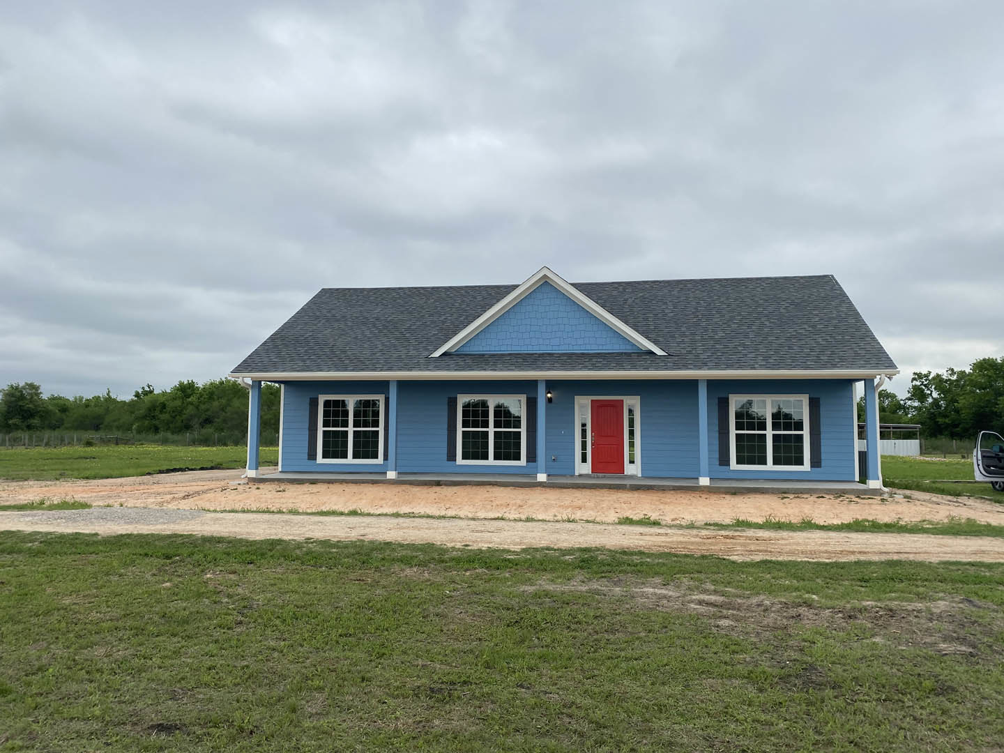 Blue siding house with white-framed windows, red front door with white trim, gray shingle roof, manicured grass lawn, and trees in the background under a partly cloudy sky