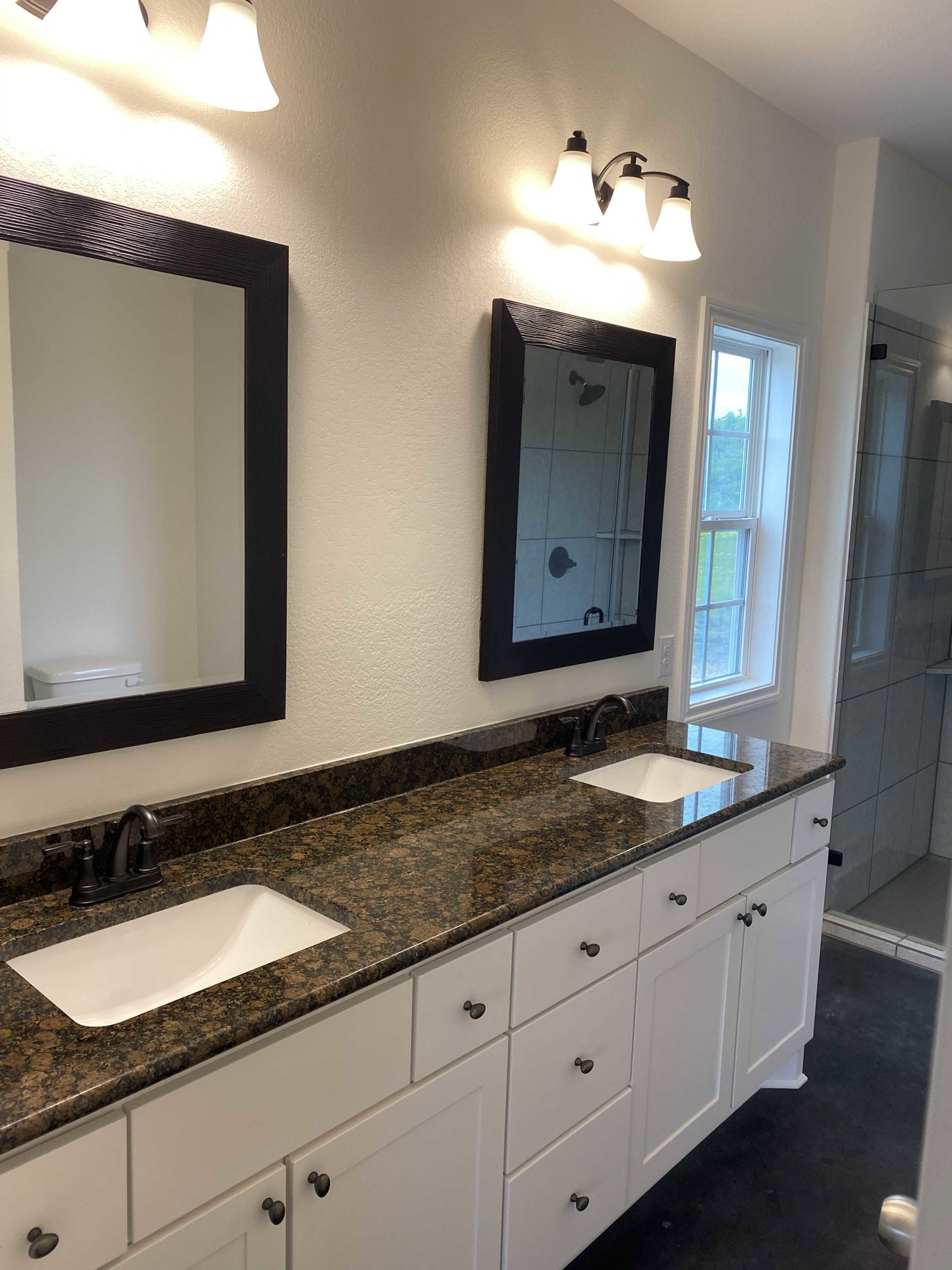 Bathroom with marble countertop, dual rectangular mirrors above white sink, three-light fixture, window with white frame, white walls, black door, and visible shower head.