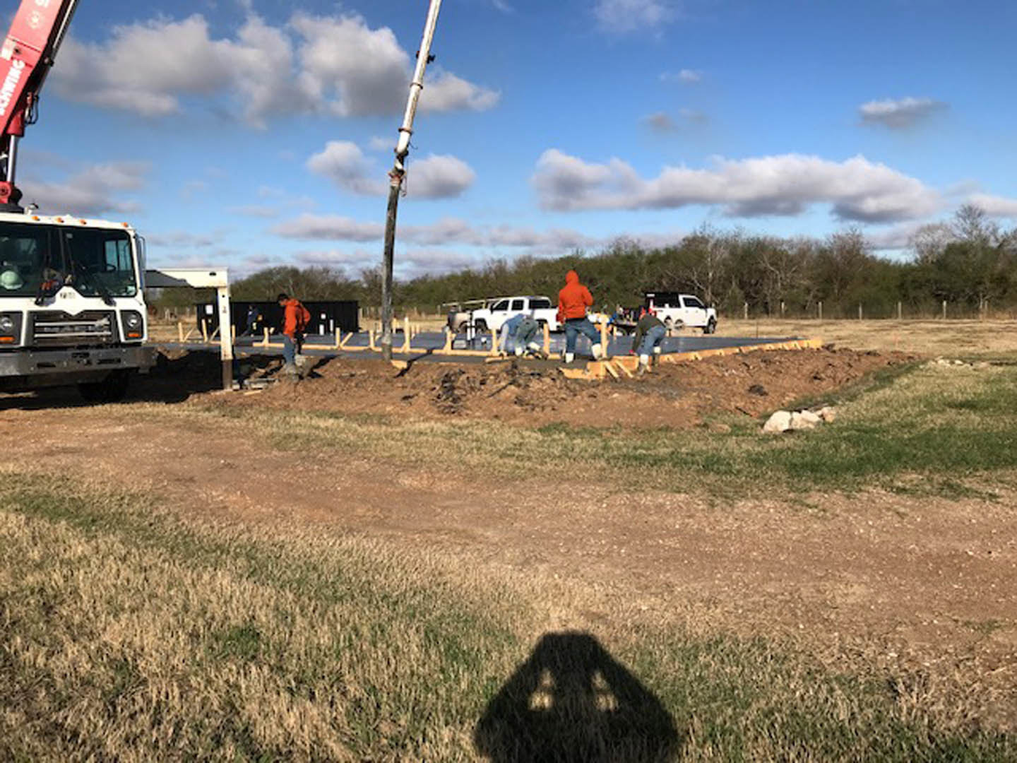 Construction workers assembling framing and materials on a grassy site beside a white truck and van, with scattered tools and vehicles under a partly cloudy sky