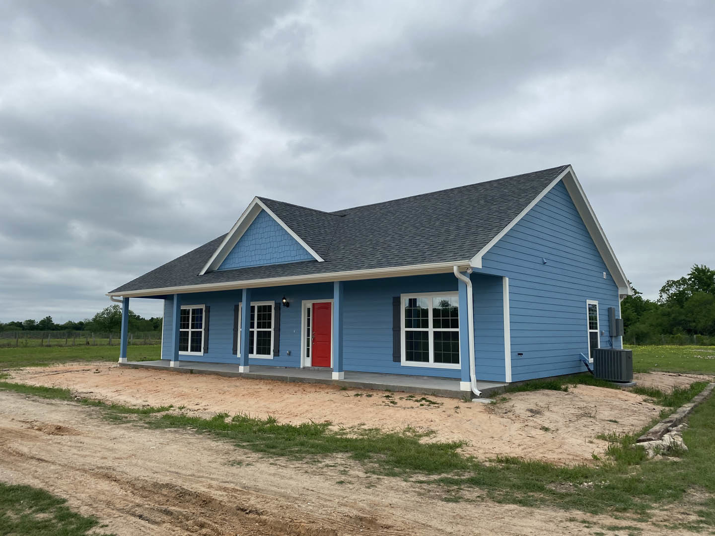 Blue siding house with white trim, red front door, and multiple windows, set beside a dirt road under a cloudy sky.