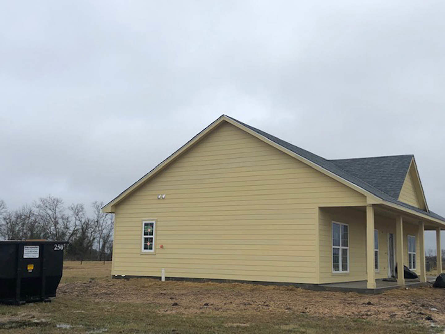 Modern custom home with light siding, white-framed windows, dark roof, and a black sign near the entrance; black dumpster with white label visible in front yard.
