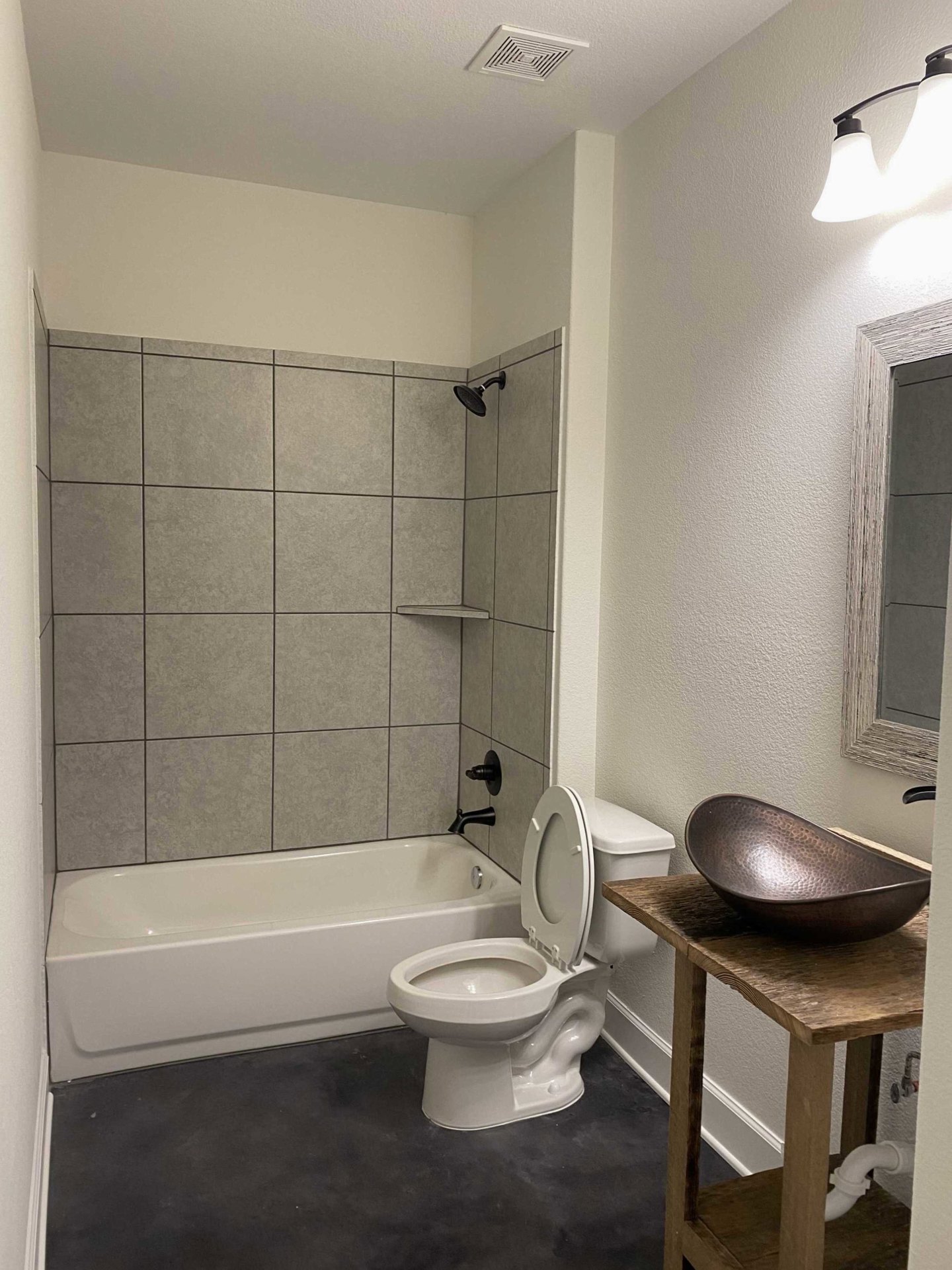 Copper vessel sink on rustic wood vanity, white toilet, freestanding bathtub, gray tile walls, modern light fixture above.