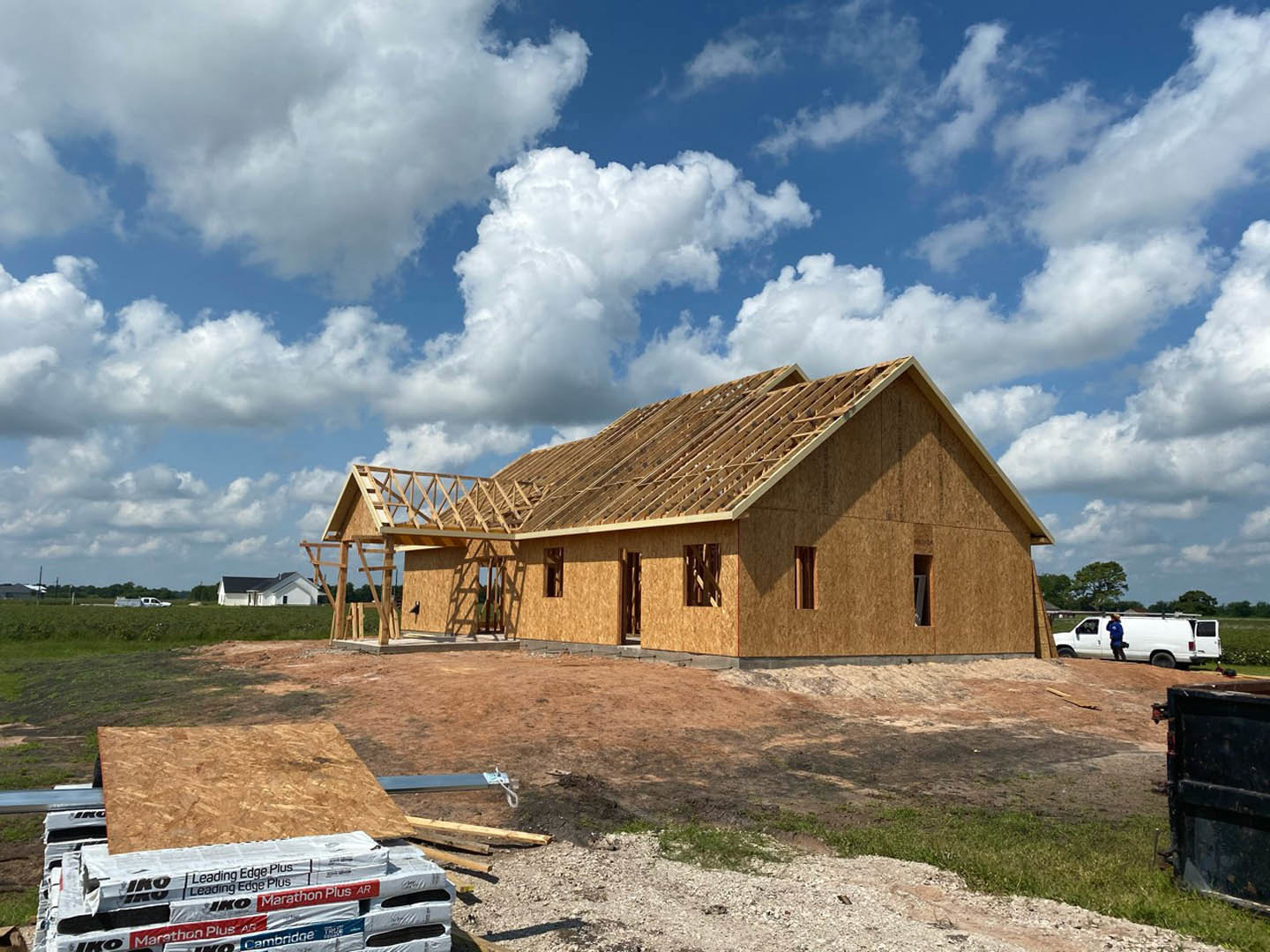 Partially built house with exposed framing and roof trusses, surrounded by grassy field, white van parked nearby, construction signs stacked in foreground, blue sky with scattered