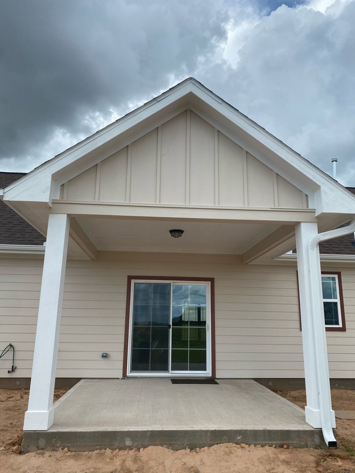 White siding house with covered porch, sliding glass door with white frame, metal railing on concrete slab, large windows, gabled roof under partly cloudy sky