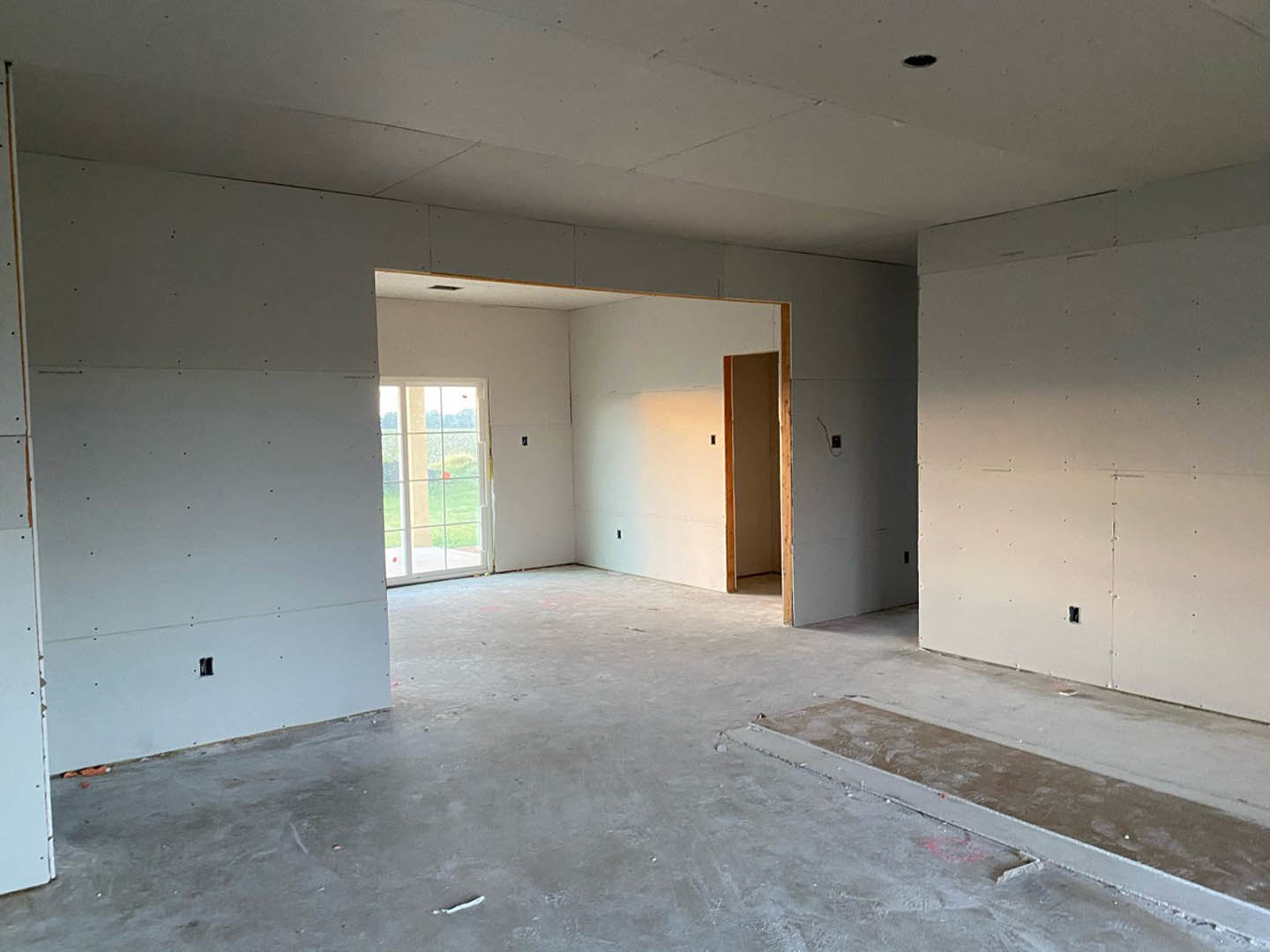 Concrete floor and white plaster walls with wooden framing, open door leading outside, window showing grassy yard with red flower, unfinished wall with visible holes