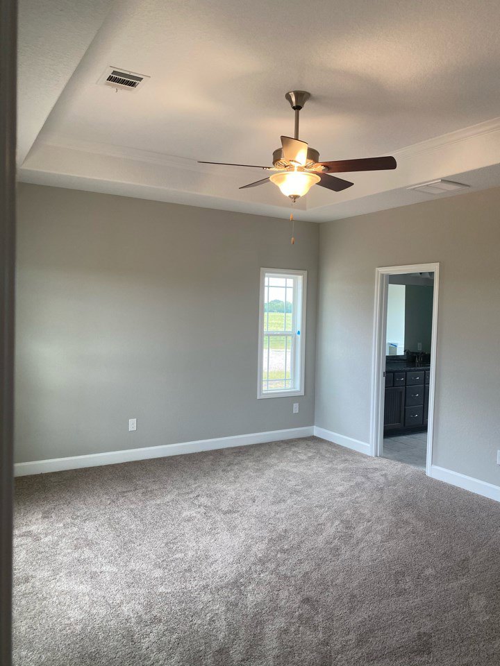 Carpeted room with white walls, ceiling fan with light fixture, window overlooking green field, doorway leading to kitchen